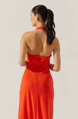 A woman is wearing a strapless, backless red dress with a fitted bodice and a high waistline. The dress has a halter neck strap that ties at the back of her neck. The skirt is long and flowing, made of a shiny, satin-like fabric. The woman has dark hair styled in a high ponytail and is wearing gold hoop earrings.