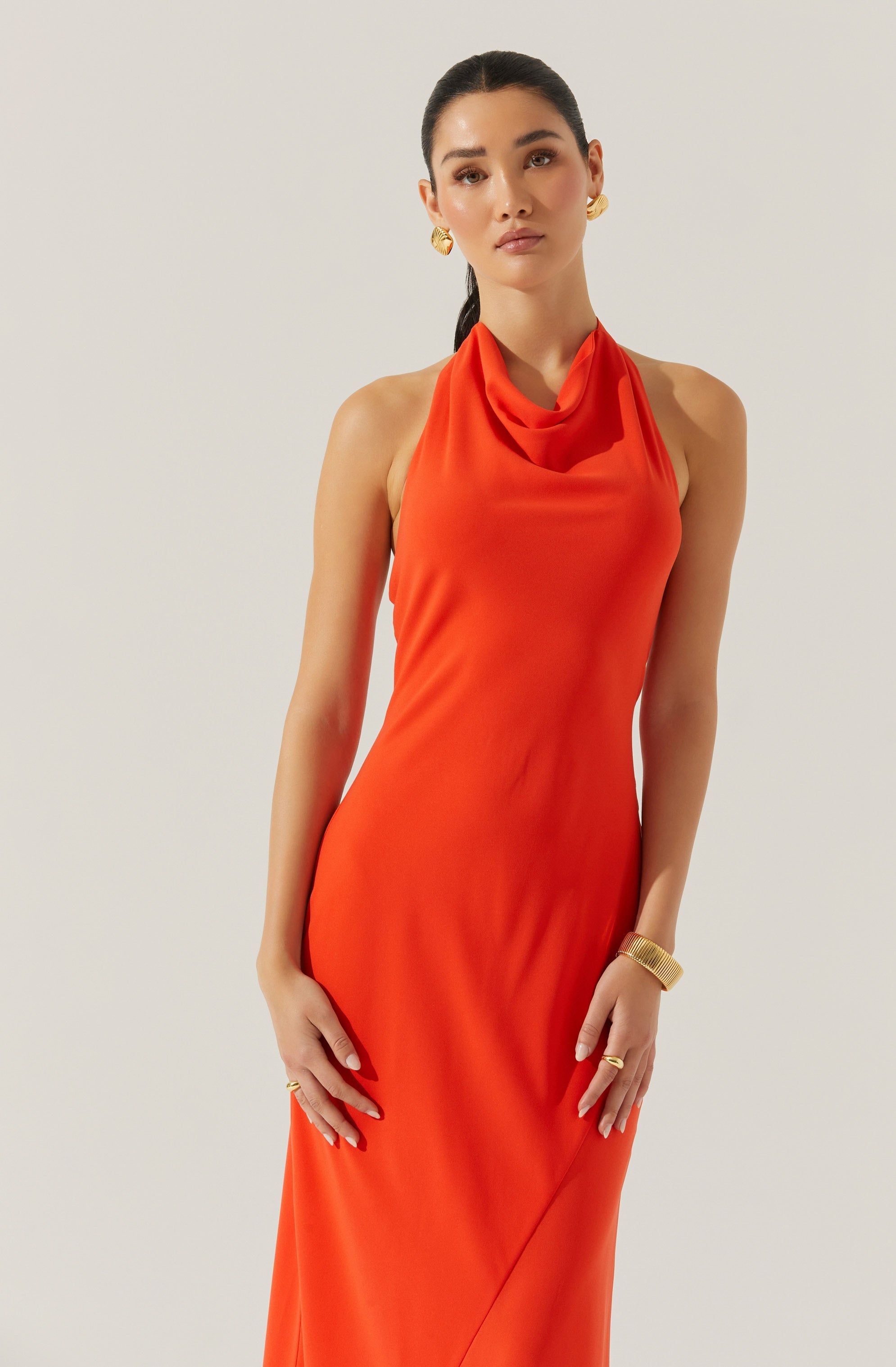 A woman is wearing a sleeveless, floor-length, bright orange satin gown with a draped cowl neckline.