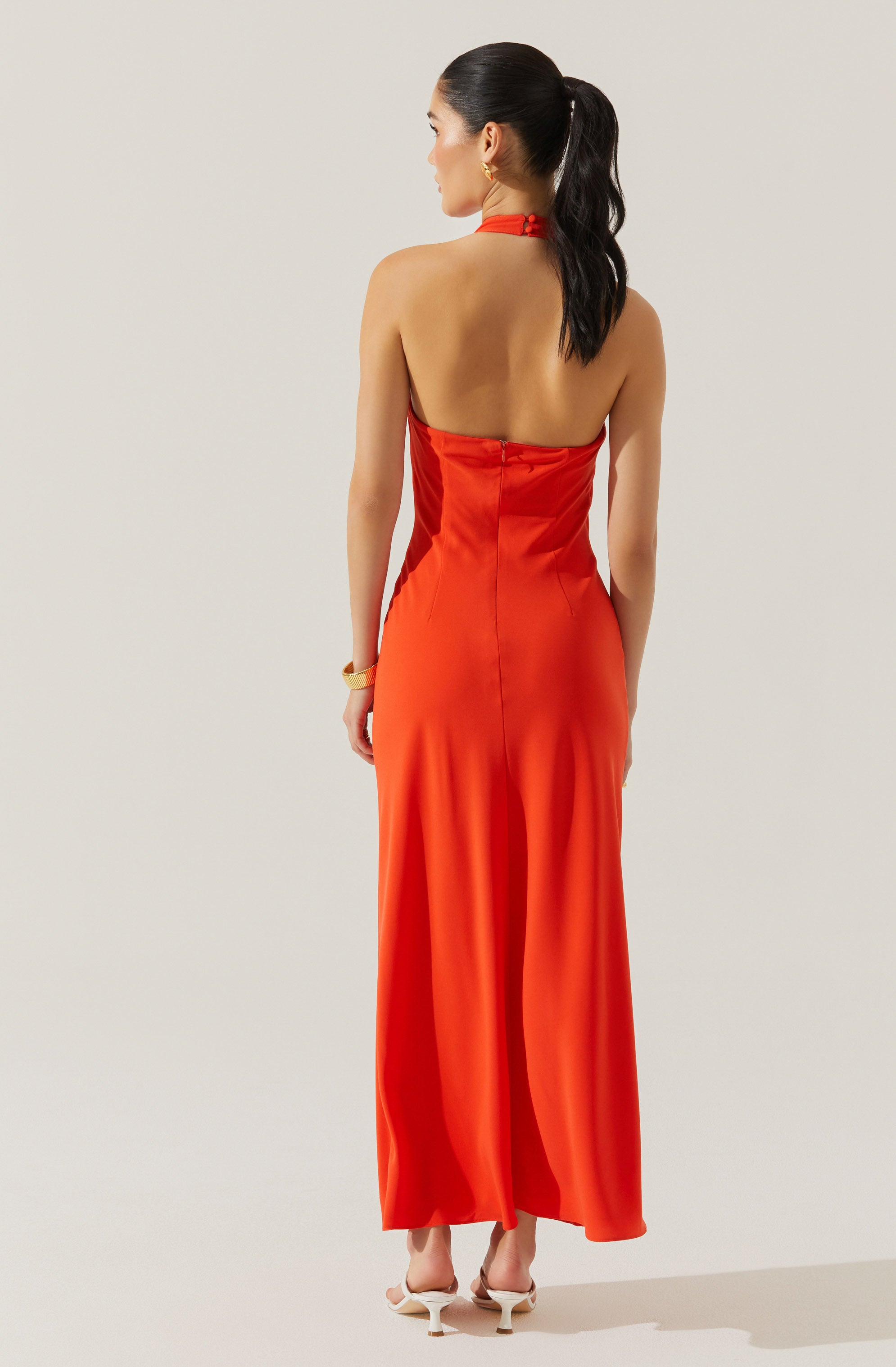 A woman is wearing a sleeveless, halter-neck, floor-length dress in a vibrant red color. The dress features a fitted bodice with a zipper closure at the back and flows into a loose, flowing skirt. She is also wearing white high-heeled shoes and a gold bracelet on her right wrist.