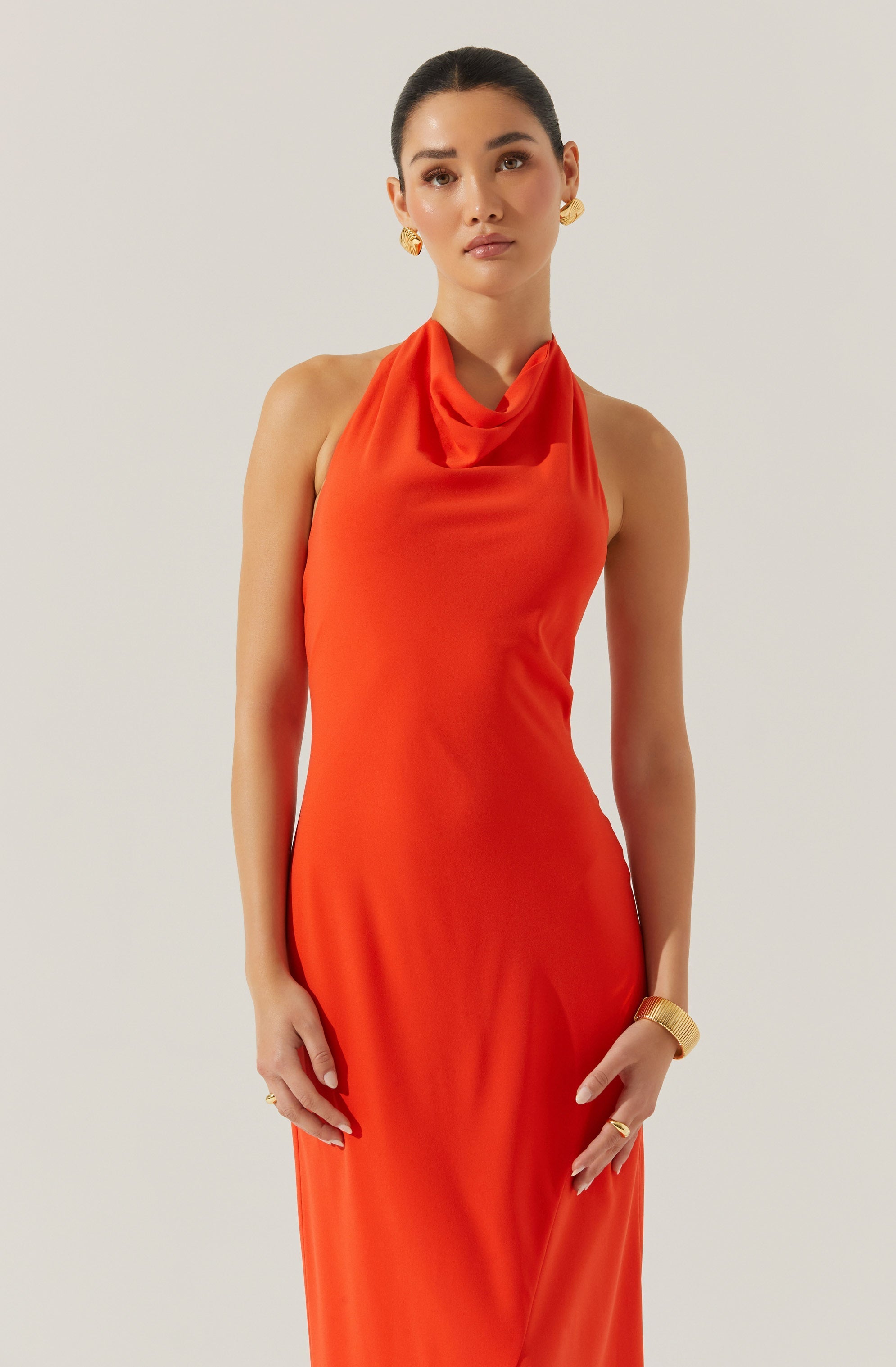 A woman is wearing a sleeveless, floor-length dress in a bright orange color. The dress features a draped neckline that creates a soft cowl effect around the collarbone. The fabric appears smooth and slightly shiny, giving the dress an elegant look. She accessorizes with gold hoop earrings, a gold bracelet, and a gold ring.