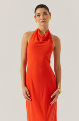 A woman is wearing a sleeveless, floor-length dress in a bright orange color. The dress features a draped neckline that creates a soft cowl effect around the collarbone. The fabric appears smooth and slightly shiny, giving the dress an elegant look. She accessorizes with gold hoop earrings, a gold bracelet, and a gold ring.