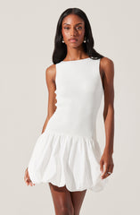 A woman is wearing a sleeveless white dress with a fitted ribbed bodice and a gathered, puffy skirt that extends just above the knees. The dress has a high, rounded neckline and features a textured fabric on the top portion, with the skirt made of a lightweight, slightly crinkled material.