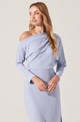 A woman is wearing a light blue off-the-shoulder dress with long sleeves. The dress has a relaxed, draped fit around the shoulders and a fitted waist. The cuffs of the sleeves are slightly darker in shade. She has long, wavy, light brown hair.
