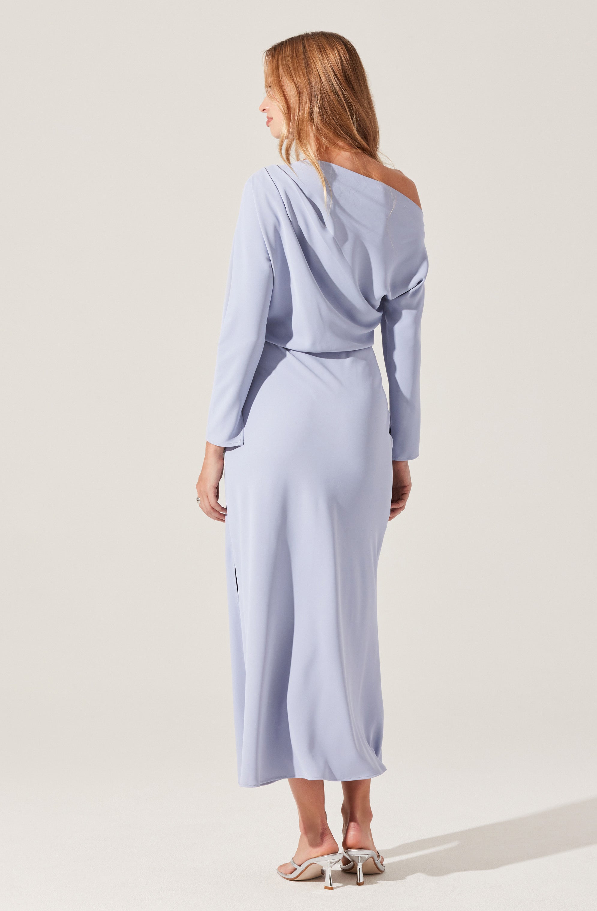 A woman is wearing a light blue, long-sleeved, off-the-shoulder dress with a loose, draped top and a fitted waist. The dress has a silky, flowing fabric and reaches down to her ankles. She is wearing beige high-heeled sandals.