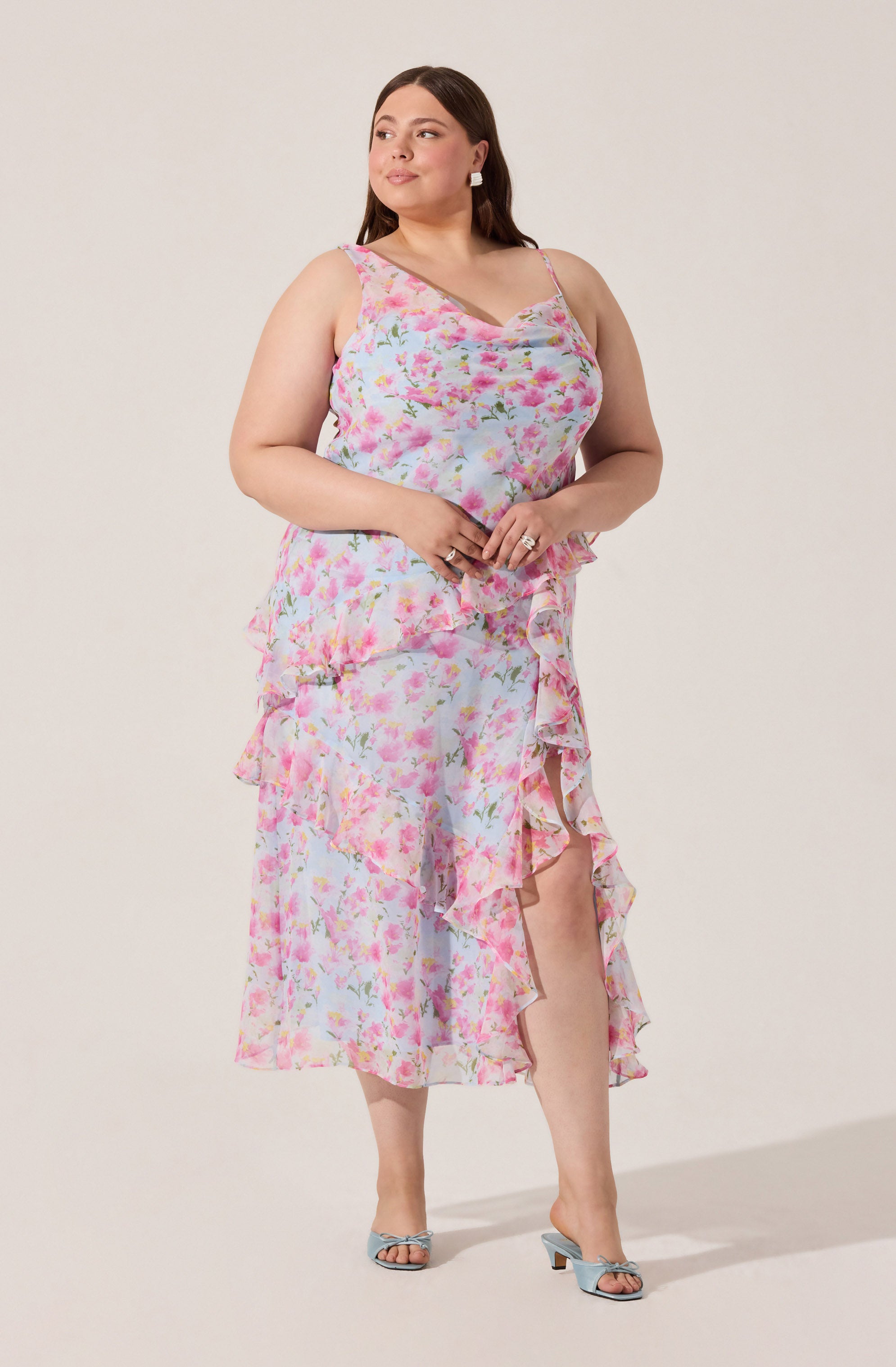 A woman is wearing a sleeveless, floral dress with a pink, white, and green pattern. The dress features thin spaghetti straps, a ruffled hemline, and a high-low style with the front shorter than the back. She is also wearing light blue open-toe high heels.