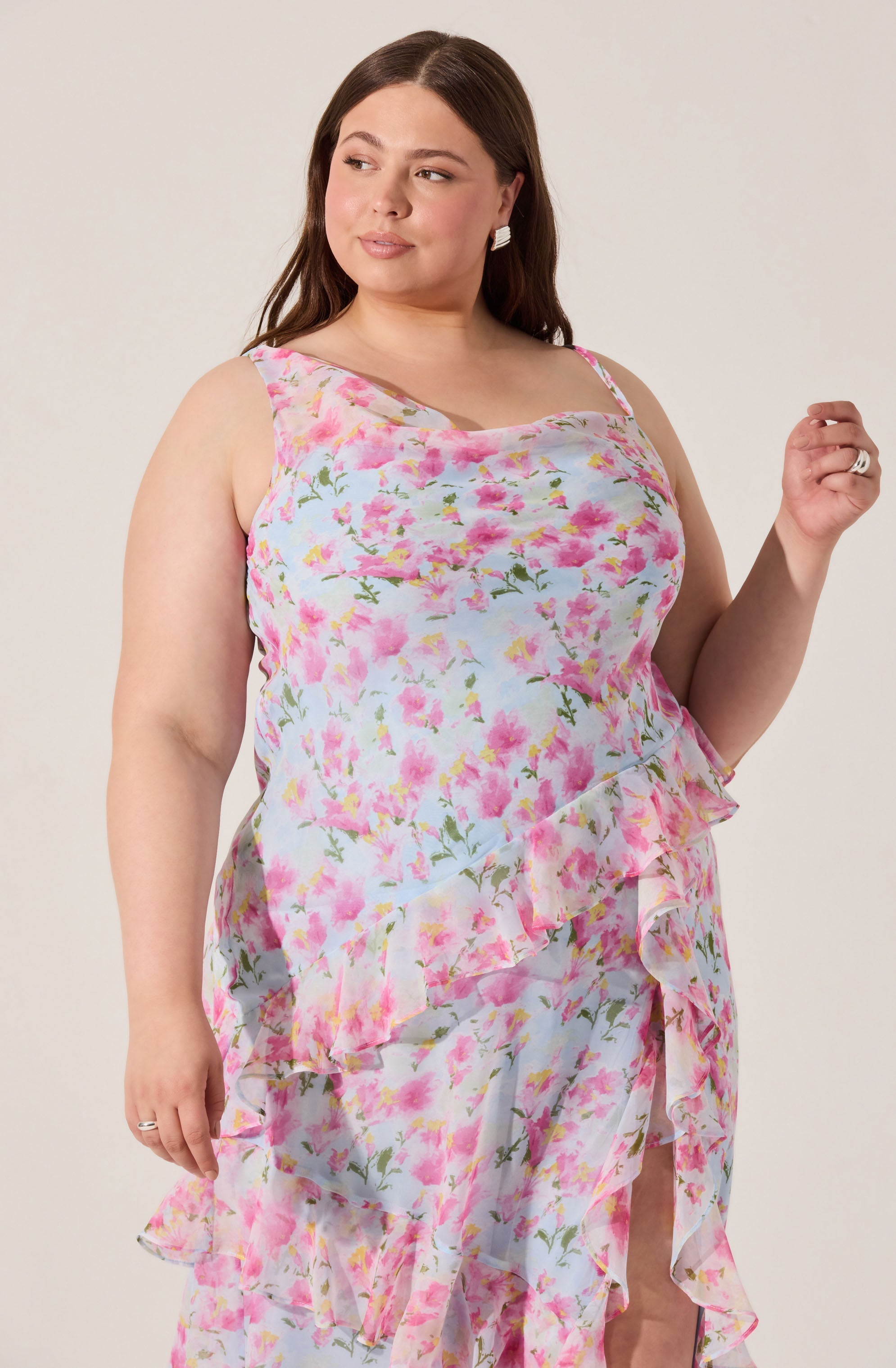 A woman wearing a sleeveless, knee-length dress with a floral pattern featuring pink, yellow, and green colors. The dress has a ruffled hemline and a sheer overlay.