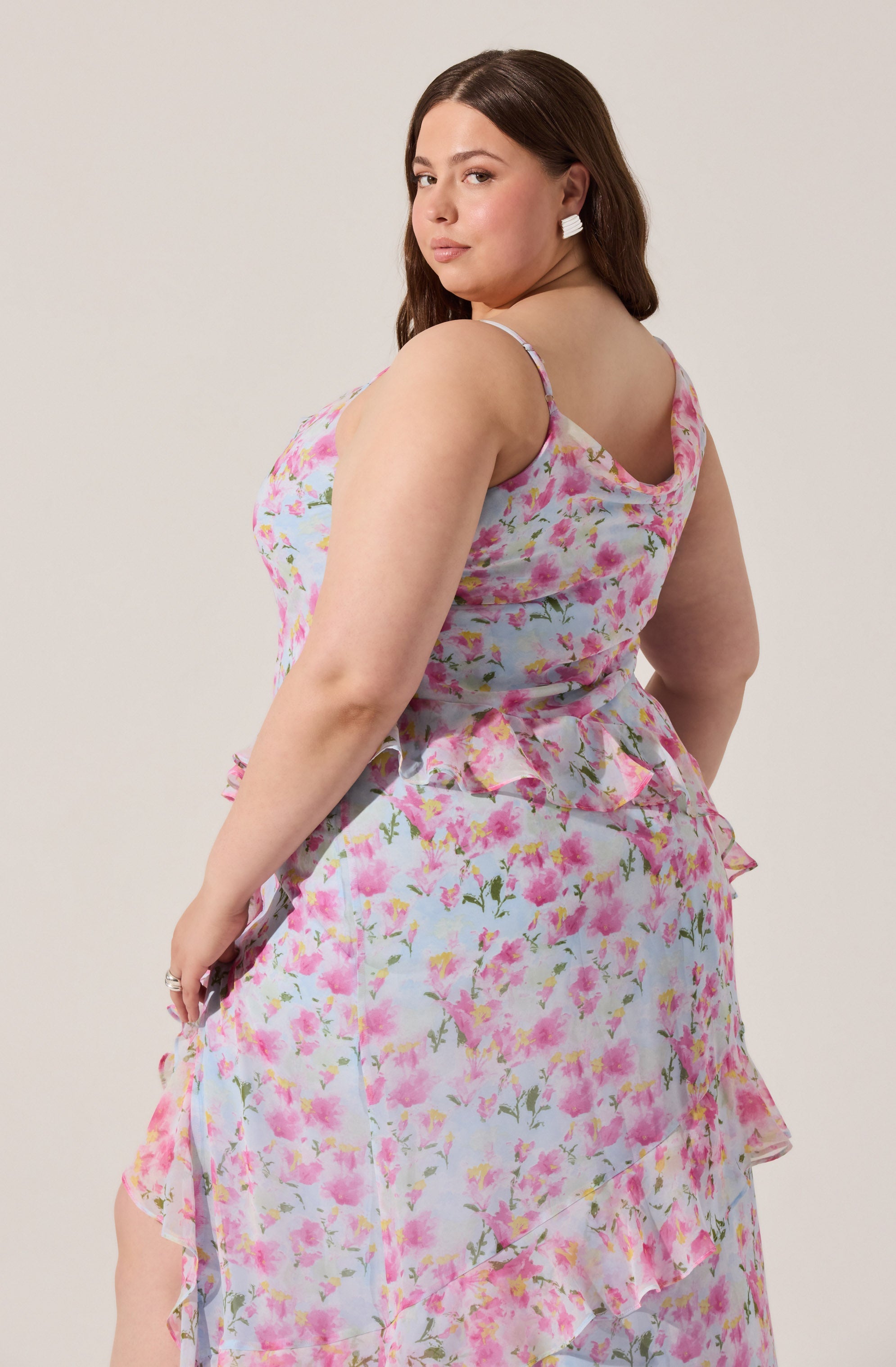 A woman is wearing a sleeveless, pastel-colored floral dress with pink, purple, and green flowers. The dress features thin straps and ruffled details on the skirt.