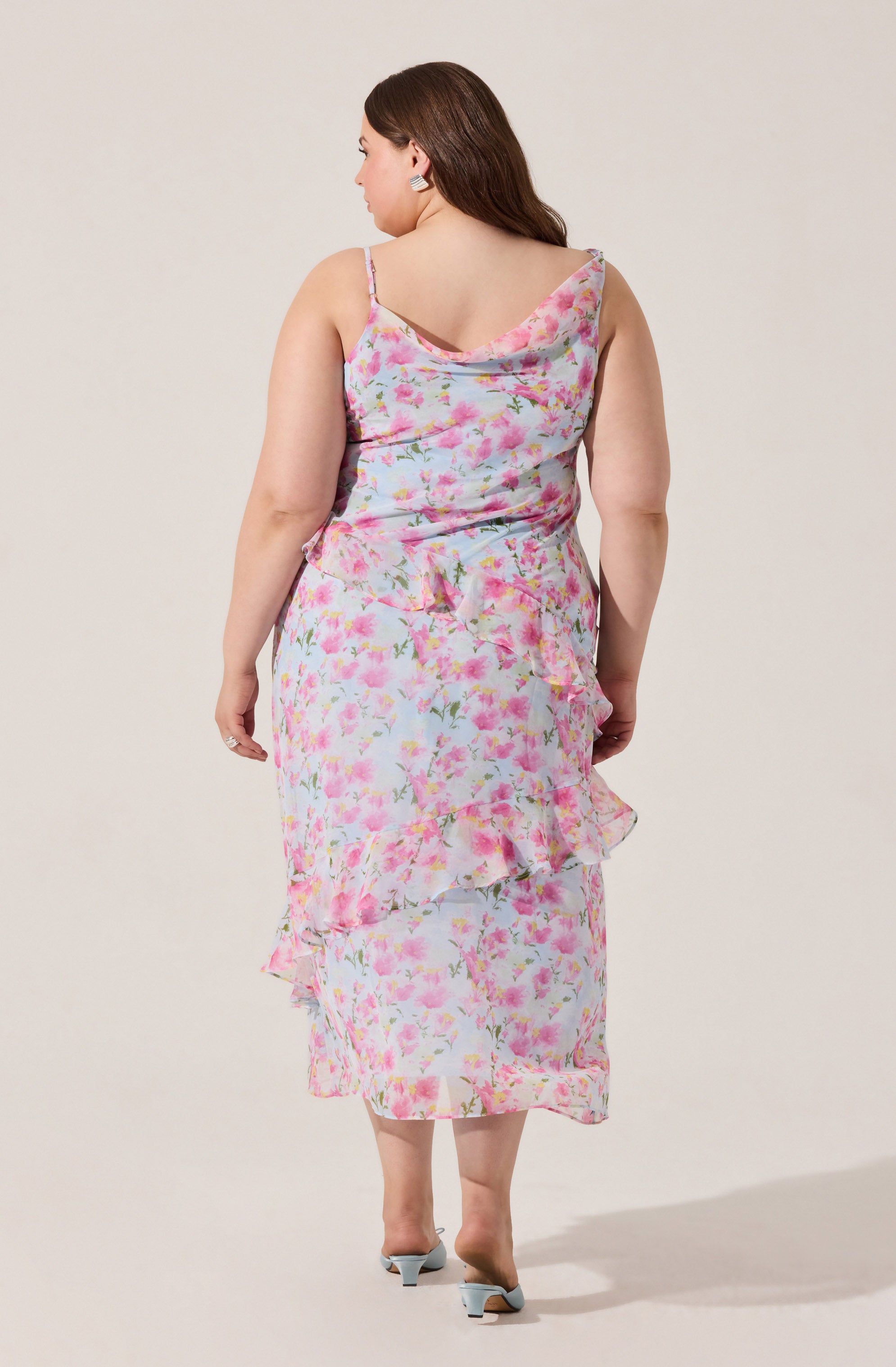A woman is wearing a sleeveless, pastel-colored, floral print dress with thin spaghetti straps. The dress features a cowl neckline at the back and ruffled layers cascading down the skirt. The fabric has a light, airy feel and the pattern consists of pink, green, and white floral designs.