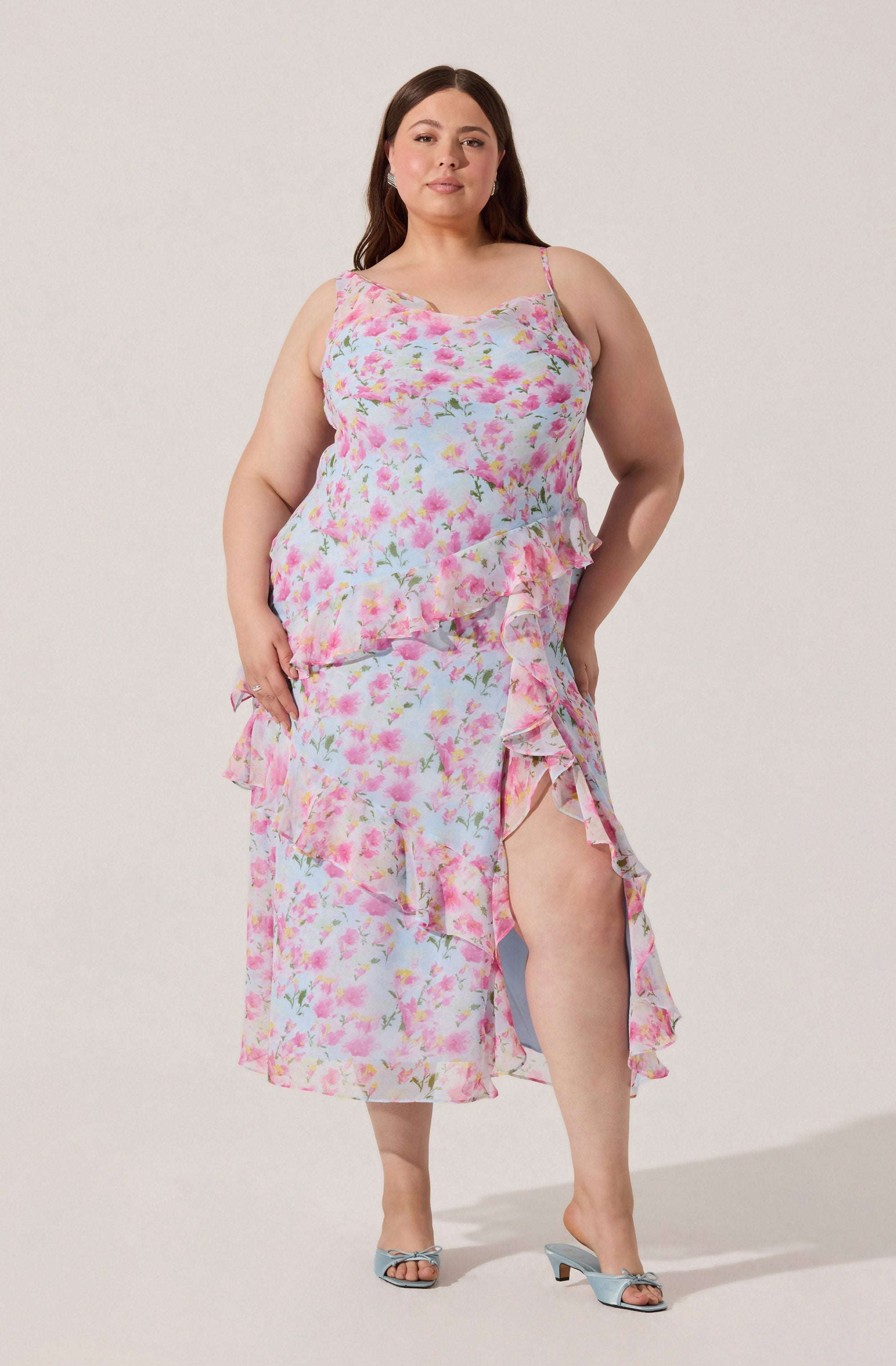 A woman is wearing a sleeveless, floral-print dress with pink, white, and green colors. The dress has thin spaghetti straps and features ruffles along the hem and side slit, revealing part of her leg. She is also wearing light blue, open-toe heeled sandals. The background is plain and light-colored.