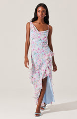 A woman is wearing a sleeveless, asymmetrical dress with spaghetti straps. The dress features a pastel watercolor floral pattern in shades of pink, blue, and green. It has ruffled details along the hemline and front slit, revealing a light blue underlayer. She is also wearing open-toe heels with a crisscross design.