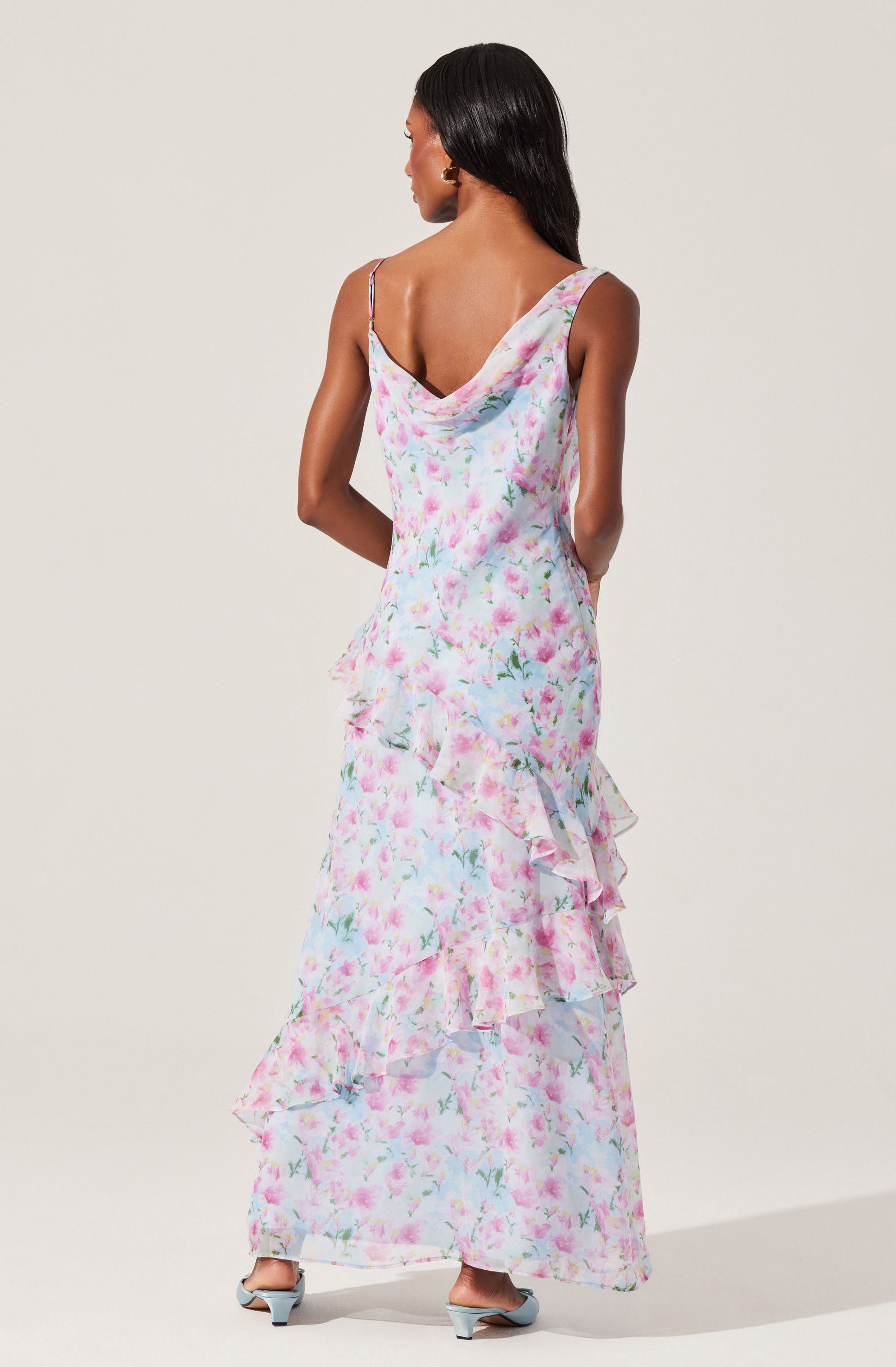 A woman is wearing a long, sleeveless dress with a floral pattern in soft pastel colors. The dress features thin spaghetti straps, a relaxed draped neckline, and a tiered, ruffled skirt that reaches the floor. The fabric appears lightweight and flowy.