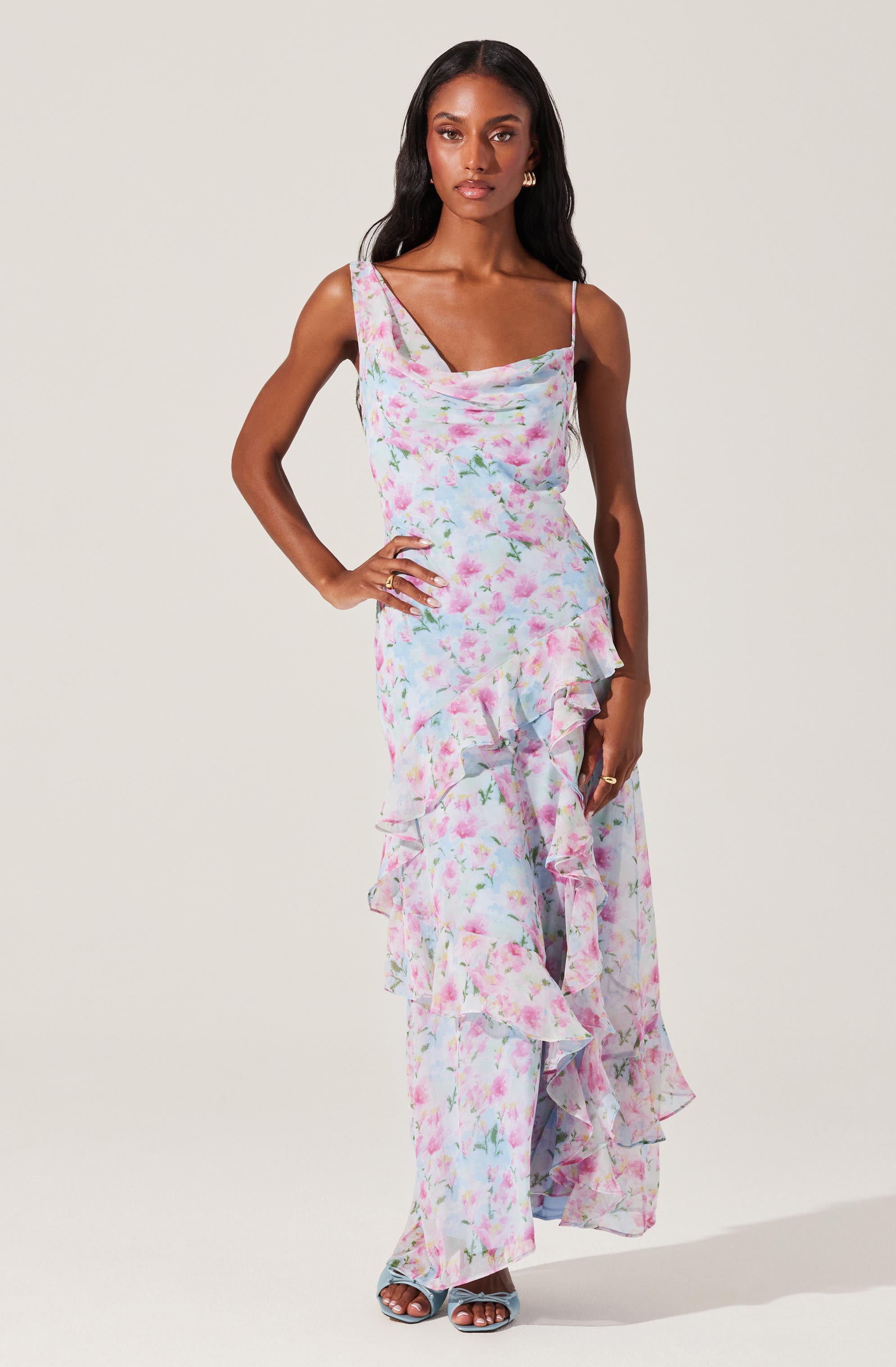 A woman is wearing a sleeveless, ankle-length dress with a pastel floral pattern. The dress features a cowl neckline, thin spaghetti straps, and ruffled details along the front and hem. She is also wearing open-toe high-heeled sandals.