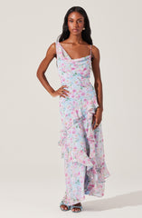 A woman is wearing a sleeveless, ankle-length dress with a pastel floral pattern. The dress features a cowl neckline, thin spaghetti straps, and ruffled details along the front and hem. She is also wearing open-toe high-heeled sandals.