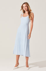 A woman is wearing a sleeveless, light blue dress with a fitted bodice and a scoop neckline. The dress features vertical pleats running from the waist down to the hem, creating a textured, flowy look that reaches mid-calf length. She is pairing the dress with light-colored, pointed-toe high heels.