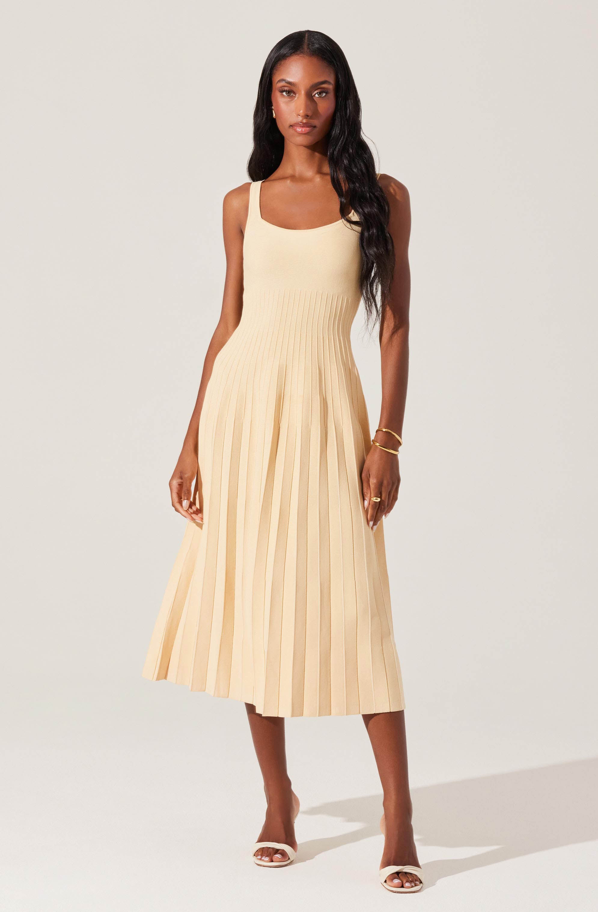 A woman is wearing a sleeveless, scoop-neck, fitted top and a high-waisted, pleated midi skirt in a light beige color. She accessorizes with a gold bracelet and ring, and is wearing open-toed heels in a matching light color.