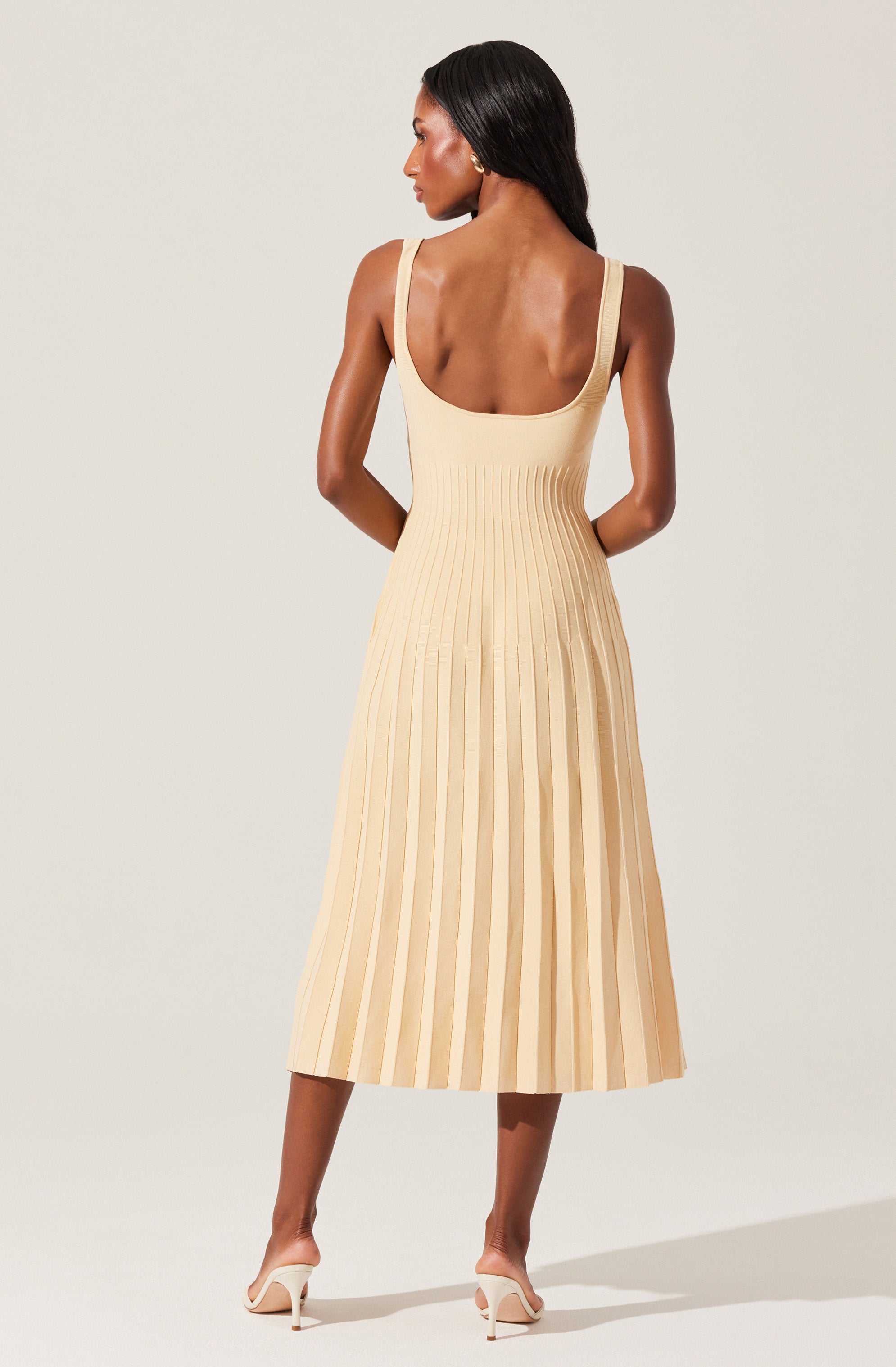 A woman is wearing a sleeveless, beige, pleated midi dress with wide shoulder straps. She is also wearing nude-colored high heels. The dress has a fitted bodice and the pleats create a flowing, A-line silhouette.