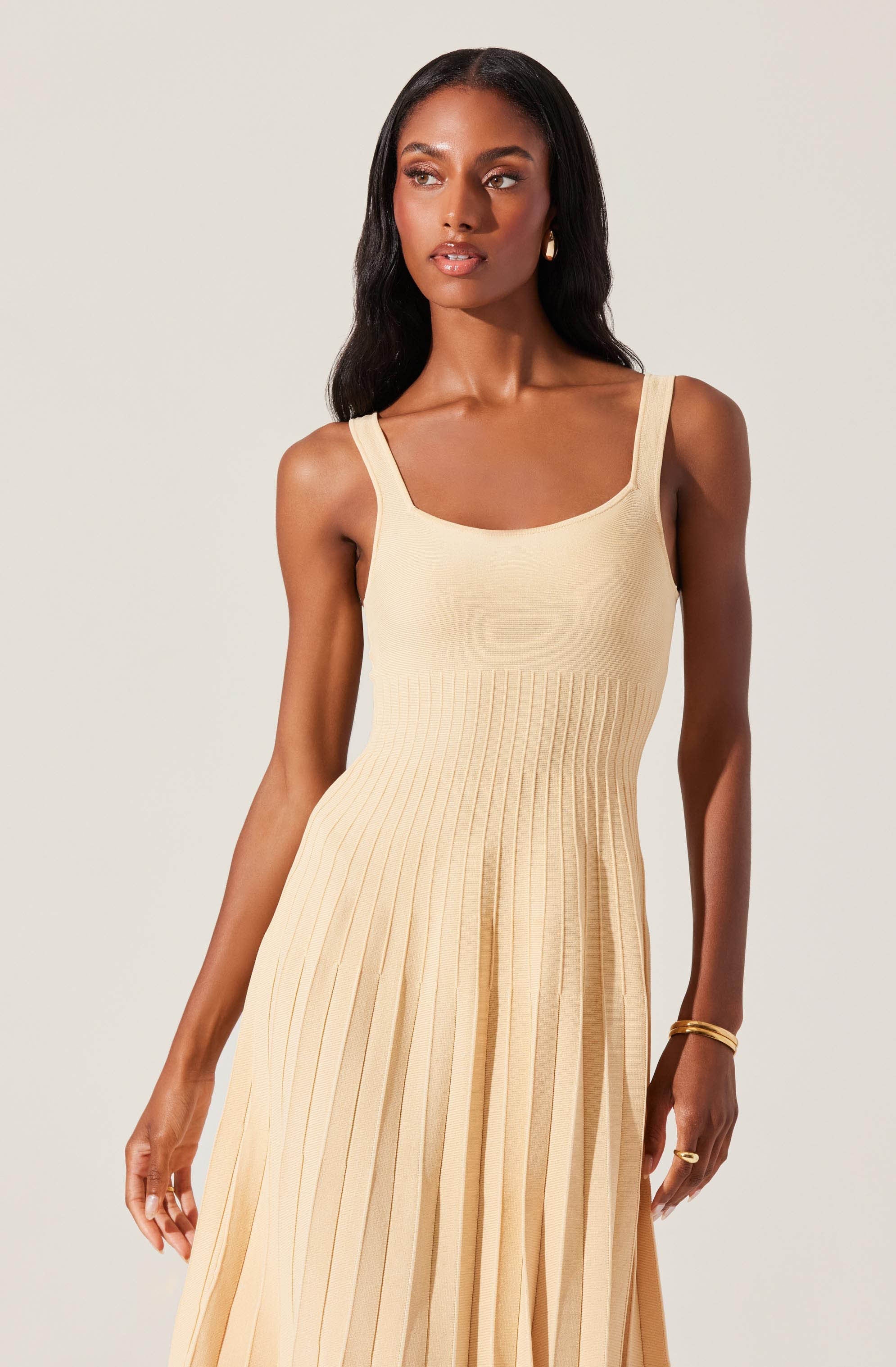 A woman is wearing a sleeveless, cream-colored dress with wide straps. The fitted bodice is smooth, and the skirt features vertical pleats that create a flowing, structured look. The dress has a simple, elegant style with a natural, light hue.