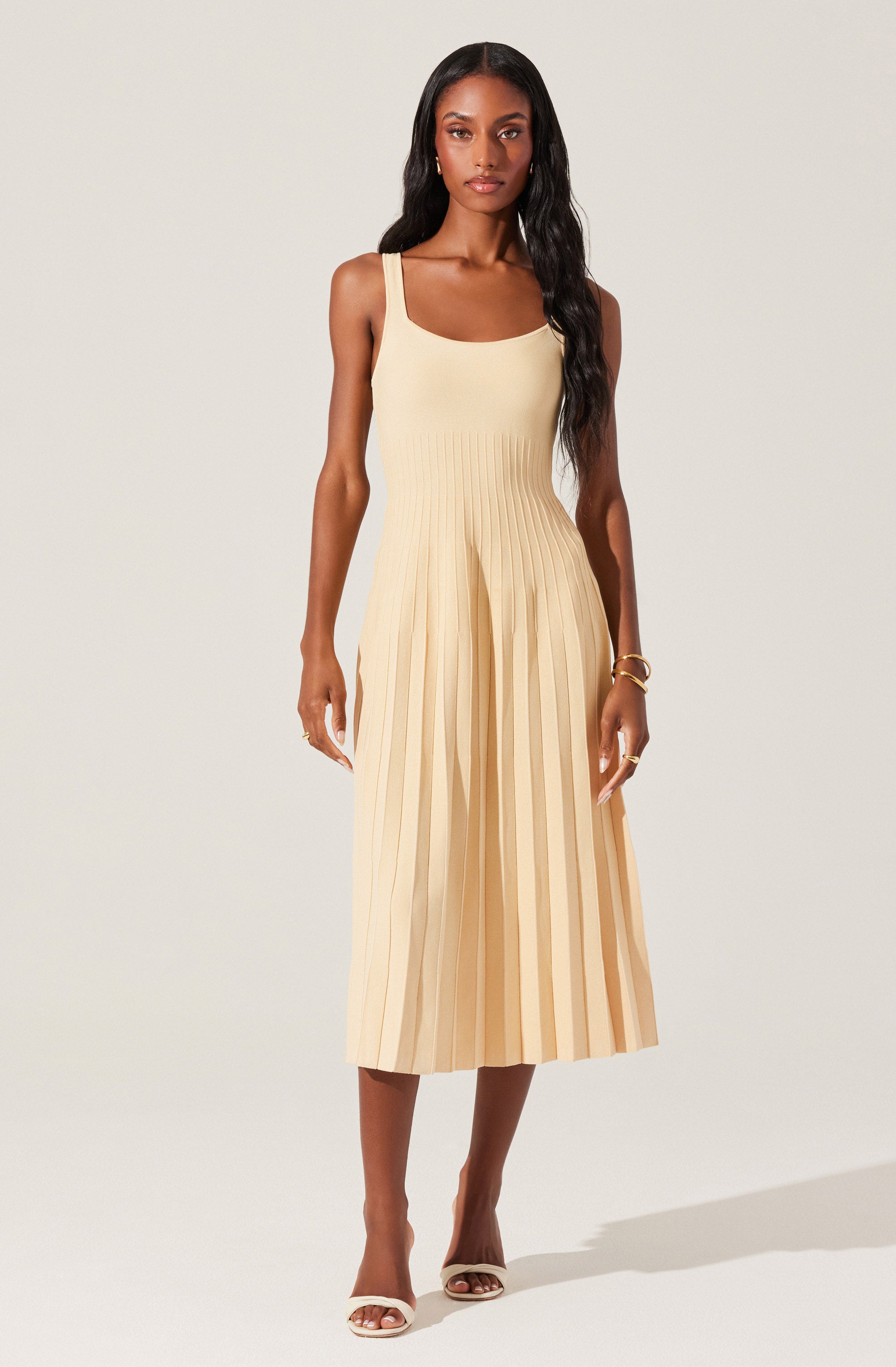 A woman is wearing a sleeveless, cream-colored dress with wide shoulder straps. The dress has a fitted bodice and features vertical pleats from the waist down, creating a flowing skirt that falls to mid-calf length. She is accessorized with gold jewelry, including bracelets and rings, and is wearing white heeled sandals.