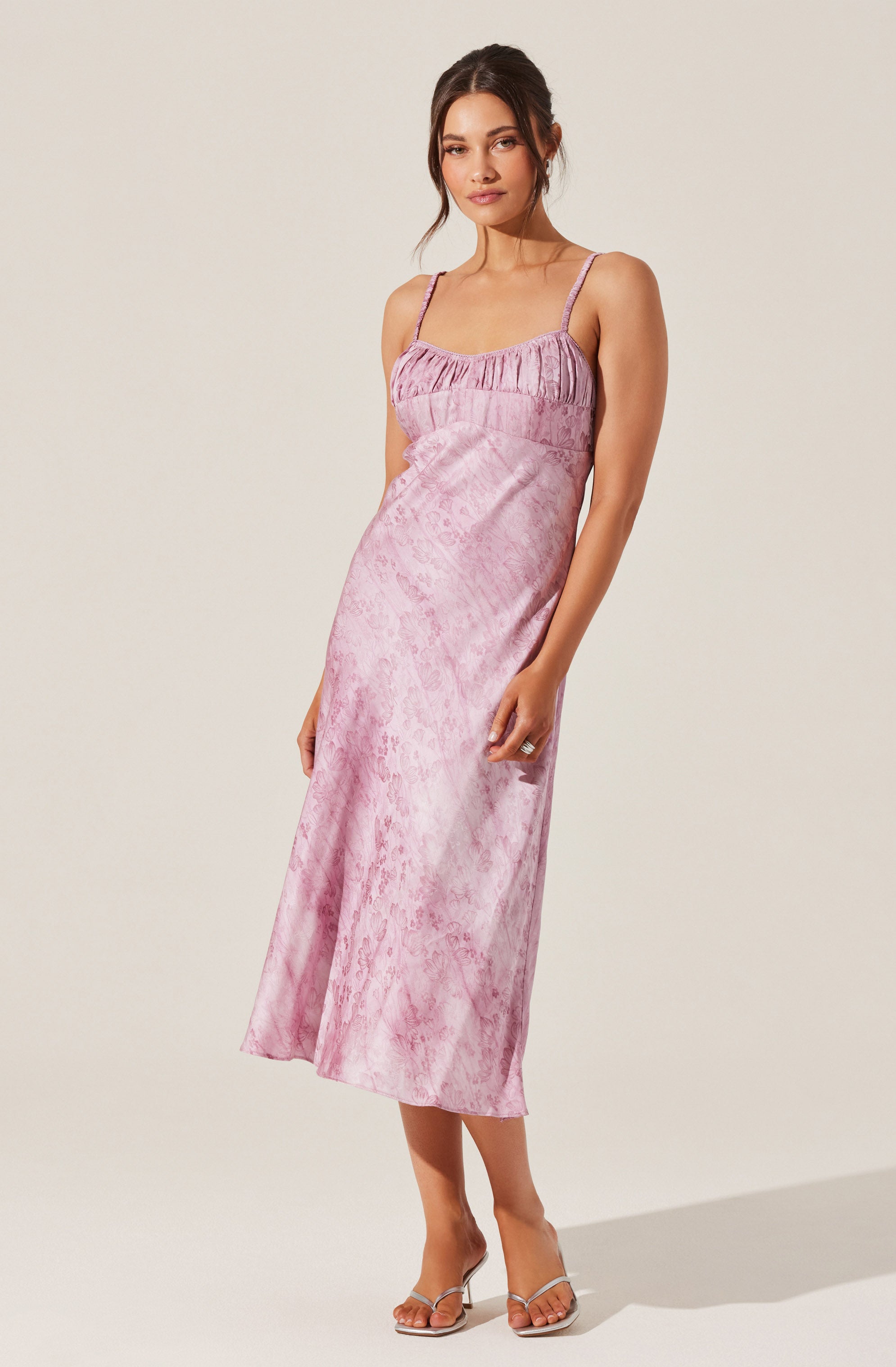 A woman is wearing a sleeveless, ankle-length dress with thin spaghetti straps. The dress has a pink, shiny, textured fabric with a subtle pattern, and features a gathered, ruffled detail at the top. She is also wearing open-toe, high-heeled sandals with clear straps.
