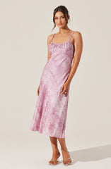 A woman is wearing a sleeveless, ankle-length dress with thin spaghetti straps. The dress has a pink, shiny, textured fabric with a subtle pattern, and features a gathered, ruffled detail at the top. She is also wearing open-toe, high-heeled sandals with clear straps.