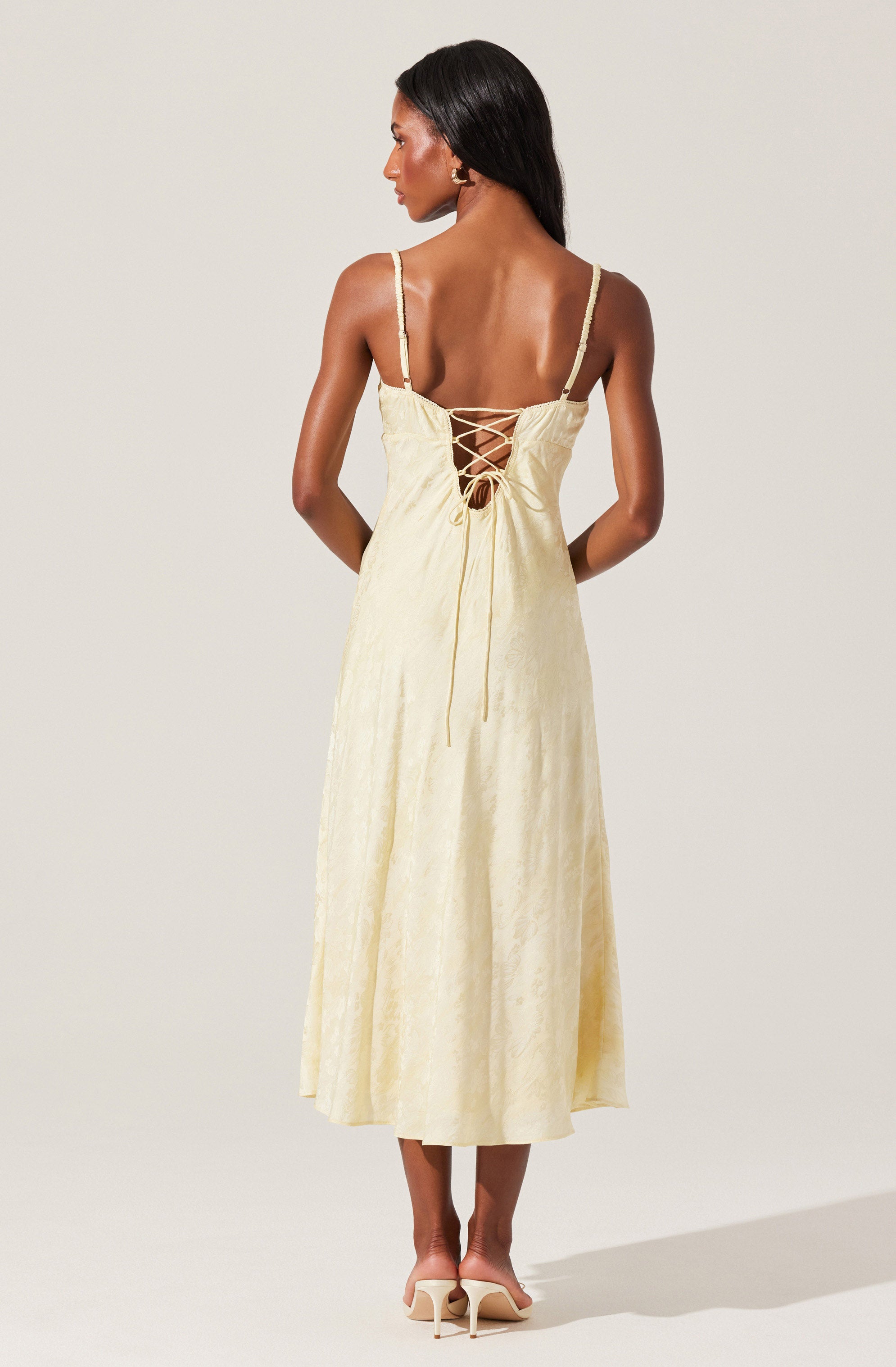 A woman is wearing a long, light yellow dress with a subtle floral pattern. The dress features spaghetti straps and a lace-up back with crisscrossing ties. The fabric appears to be lightweight and slightly textured. She is also wearing beige high-heeled shoes.