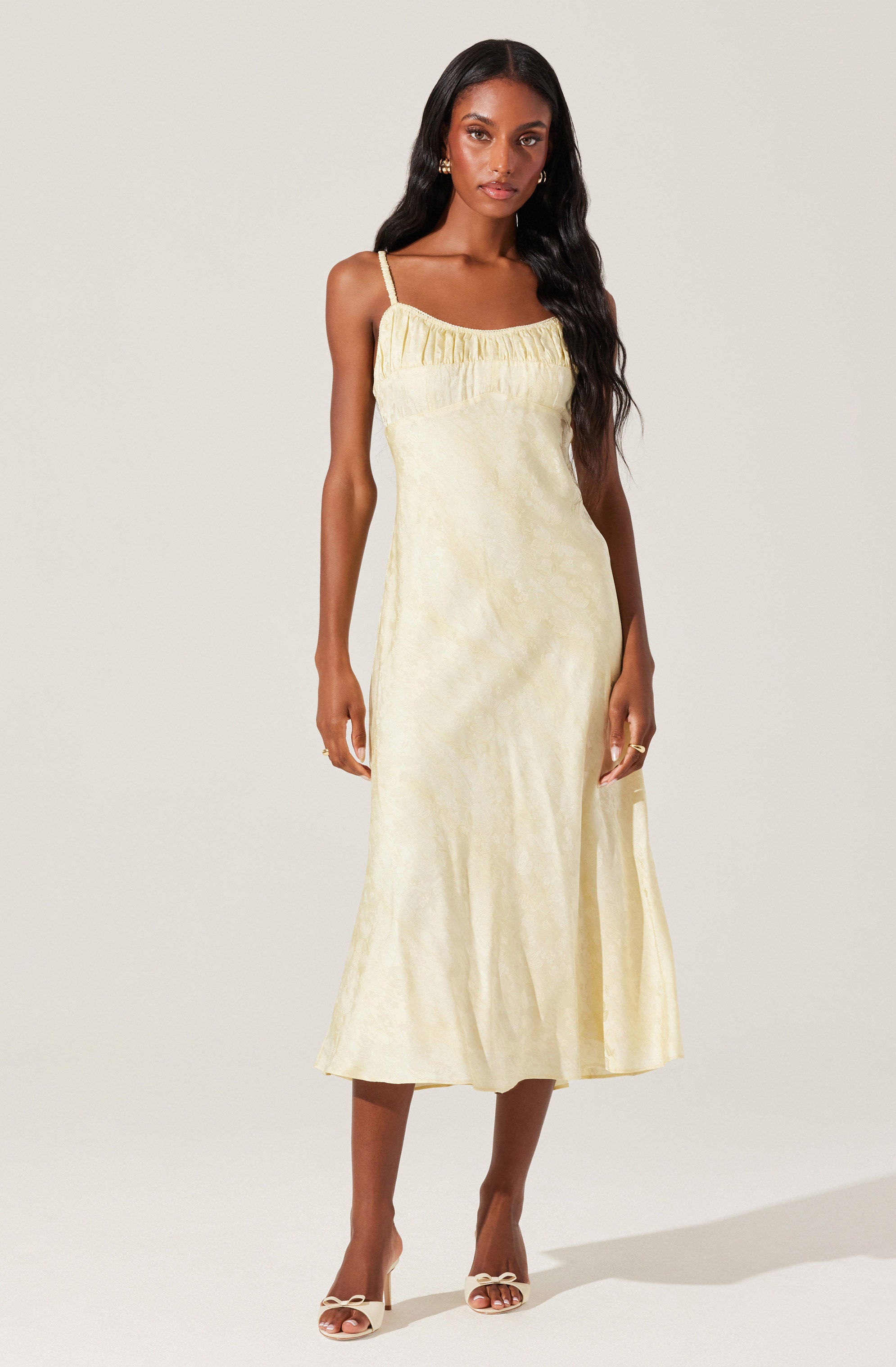 A woman is wearing a long, sleeveless, pale yellow dress with thin shoulder straps. The dress has a gathered neckline and features a subtle, textured pattern throughout. She pairs it with open-toed heels with a bow detail.
