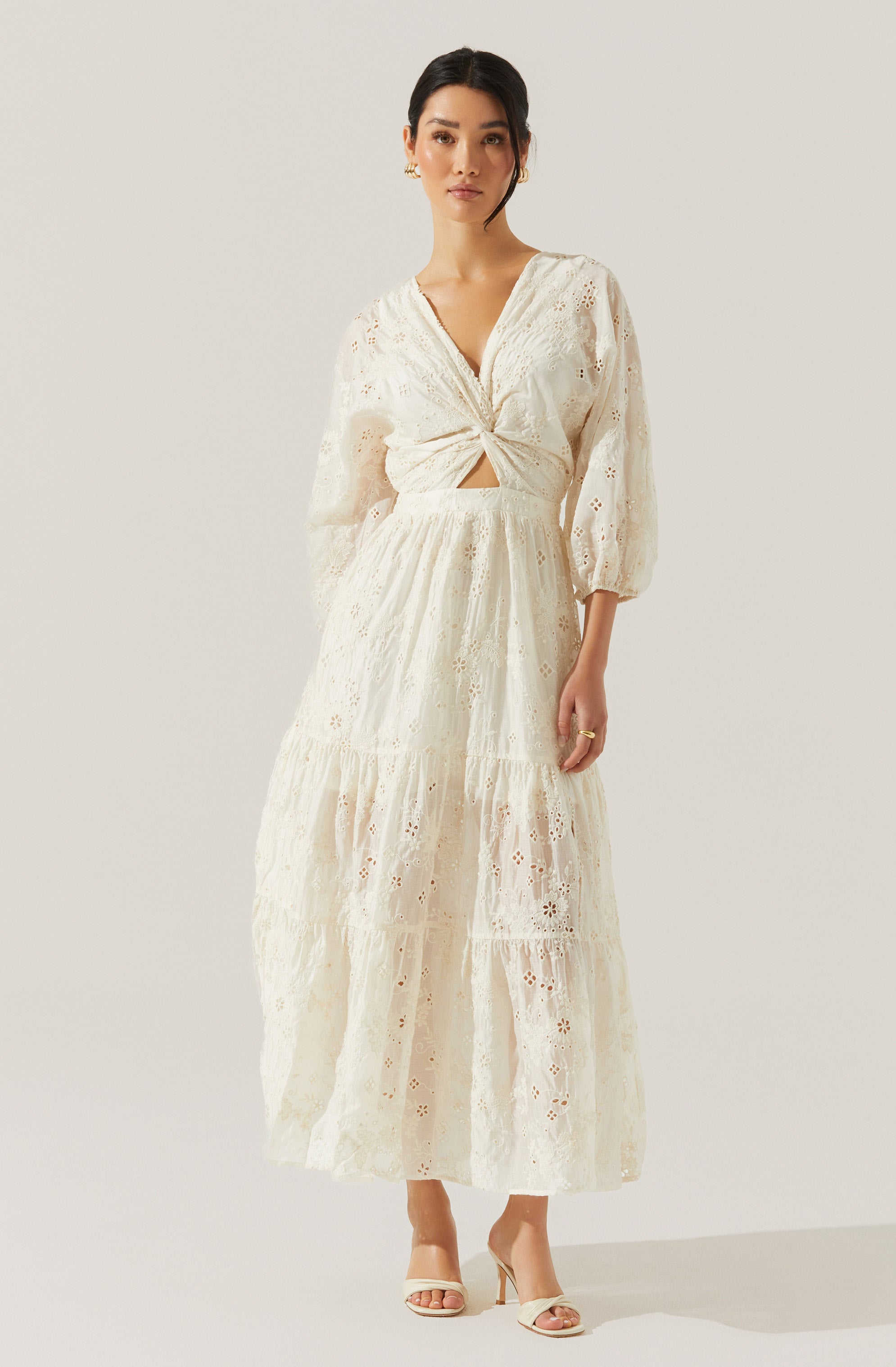 A woman is wearing a long, off-white, lace dress with eyelet embroidery. The dress features a V-neckline, a twist knot detail at the waist with a small cutout below, and voluminous three-quarter length sleeves. The skirt is tiered and flows to her ankles. She is also wearing nude-colored high-heeled sandals and gold jewelry.