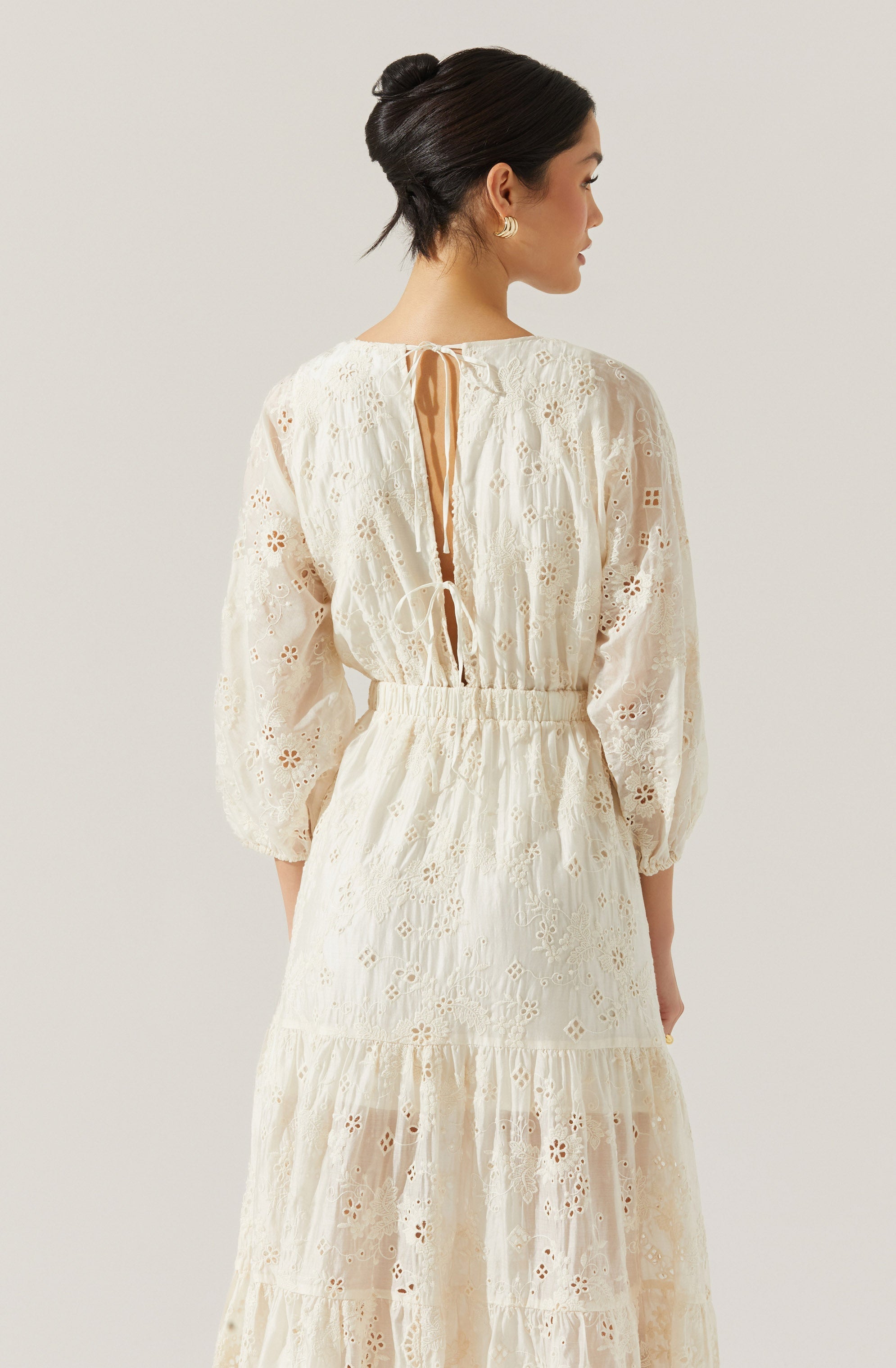 A woman is wearing a long, cream-colored dress made of eyelet lace fabric. The dress features a gathered waistband, long puffed sleeves, and a tiered skirt. The back of the dress has a keyhole opening tied with a ribbon at the neckline and a thin tie closure.
