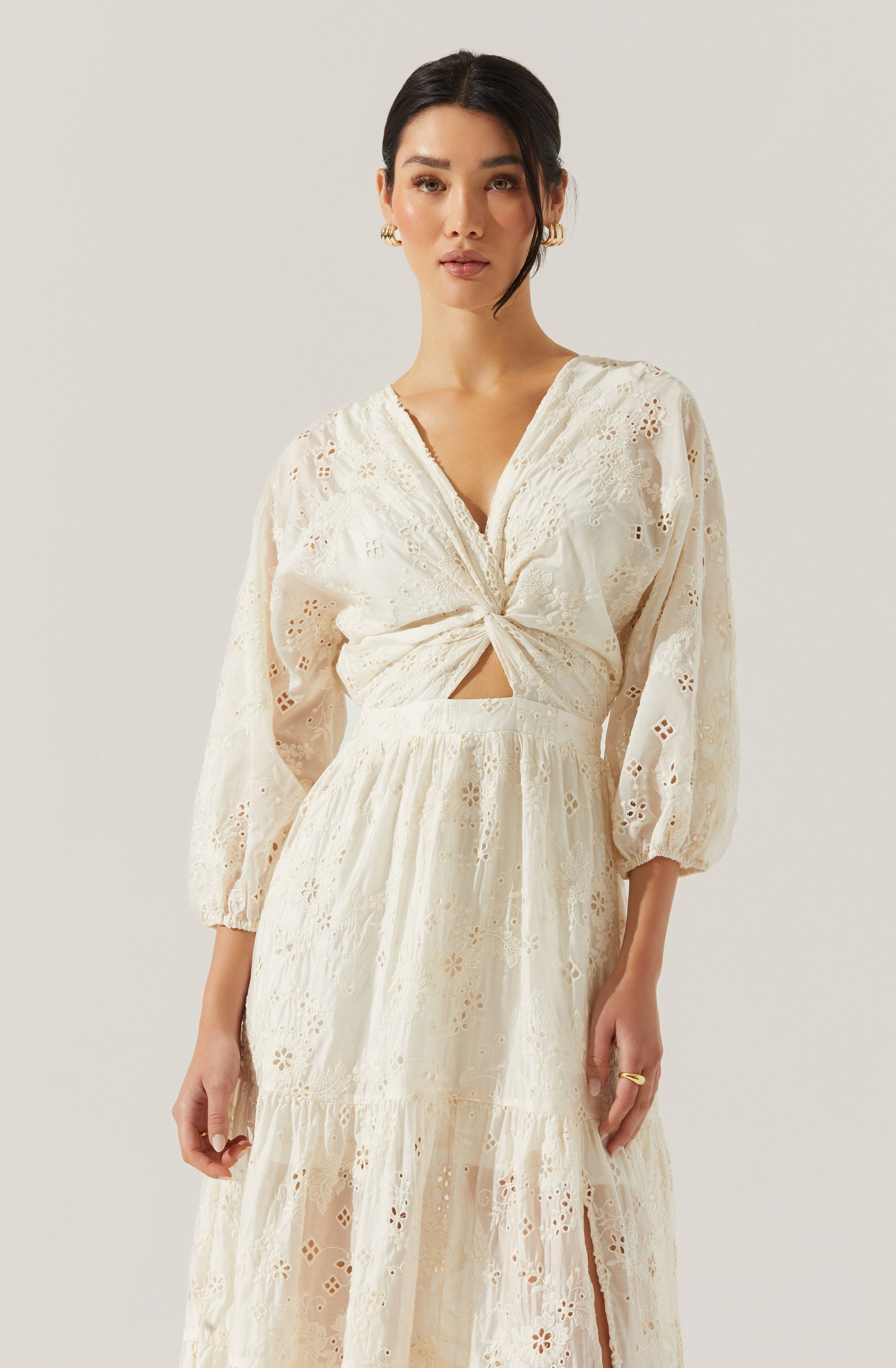 A woman is wearing a cream-colored, eyelet lace dress with intricate embroidery. The dress has long, slightly puffed sleeves gathered at the cuffs. The bodice features a V-neckline and a twist knot detail at the waist, creating a small cut-out below the knot. The skirt is flowing with a tiered design and slit detail at the front.