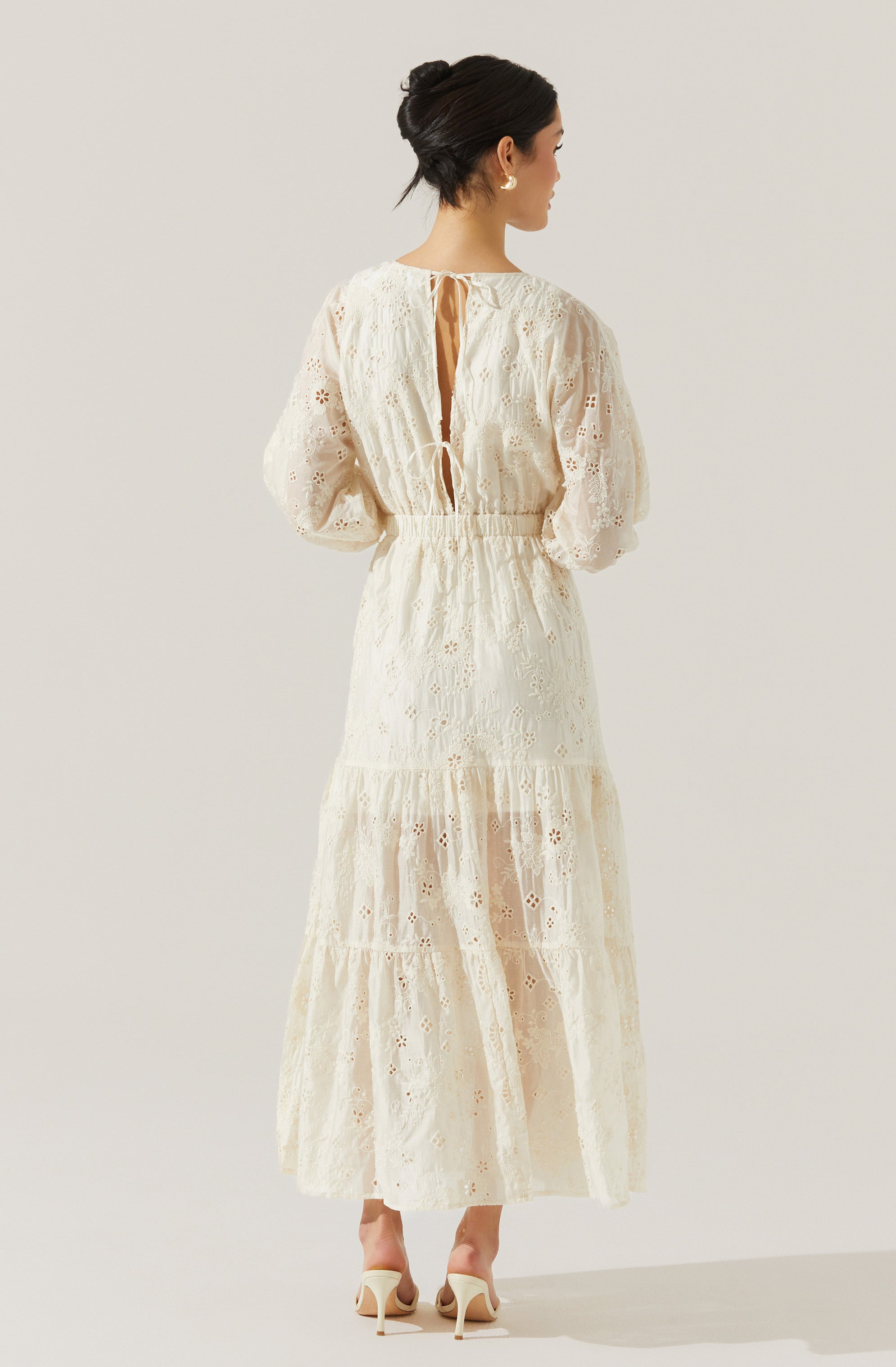 A woman is wearing an elegant, long, cream-colored dress made of eyelet lace fabric. The dress has long, puffed sleeves and a high waist. The back features a keyhole opening with a small tie at the top and an elasticated waist. The skirt is tiered, flowing down to her ankles, with intricate cut-out patterns throughout.