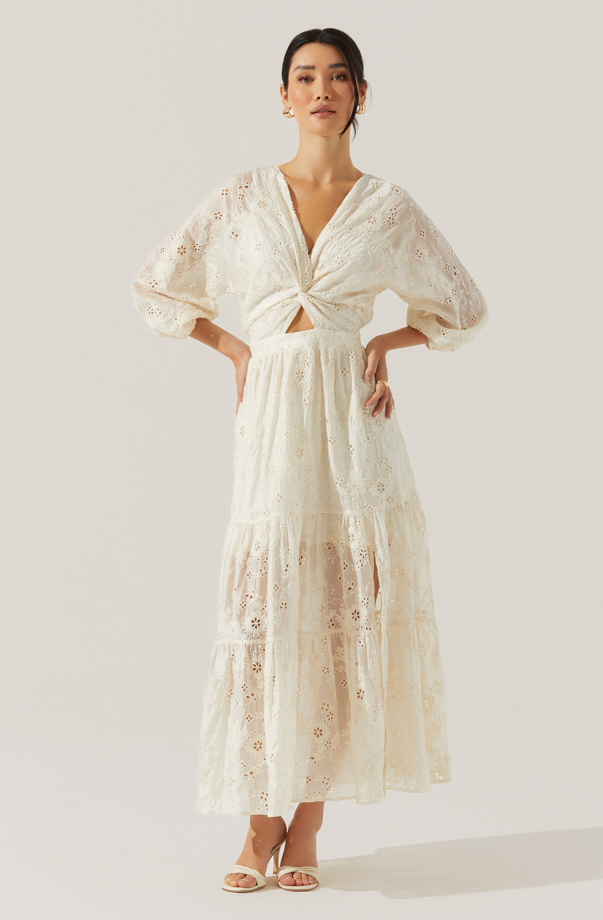 A woman is wearing a white, two-piece lace outfit with intricate eyelet embroidery. The top has long, puffed sleeves and a V-neckline that ties in a knot at the front, creating a cut-out detail at the waist. The skirt is a high-waisted, tiered maxi with a sheer lace overlay. She is also wearing open-toe, nude high-heeled sandals.