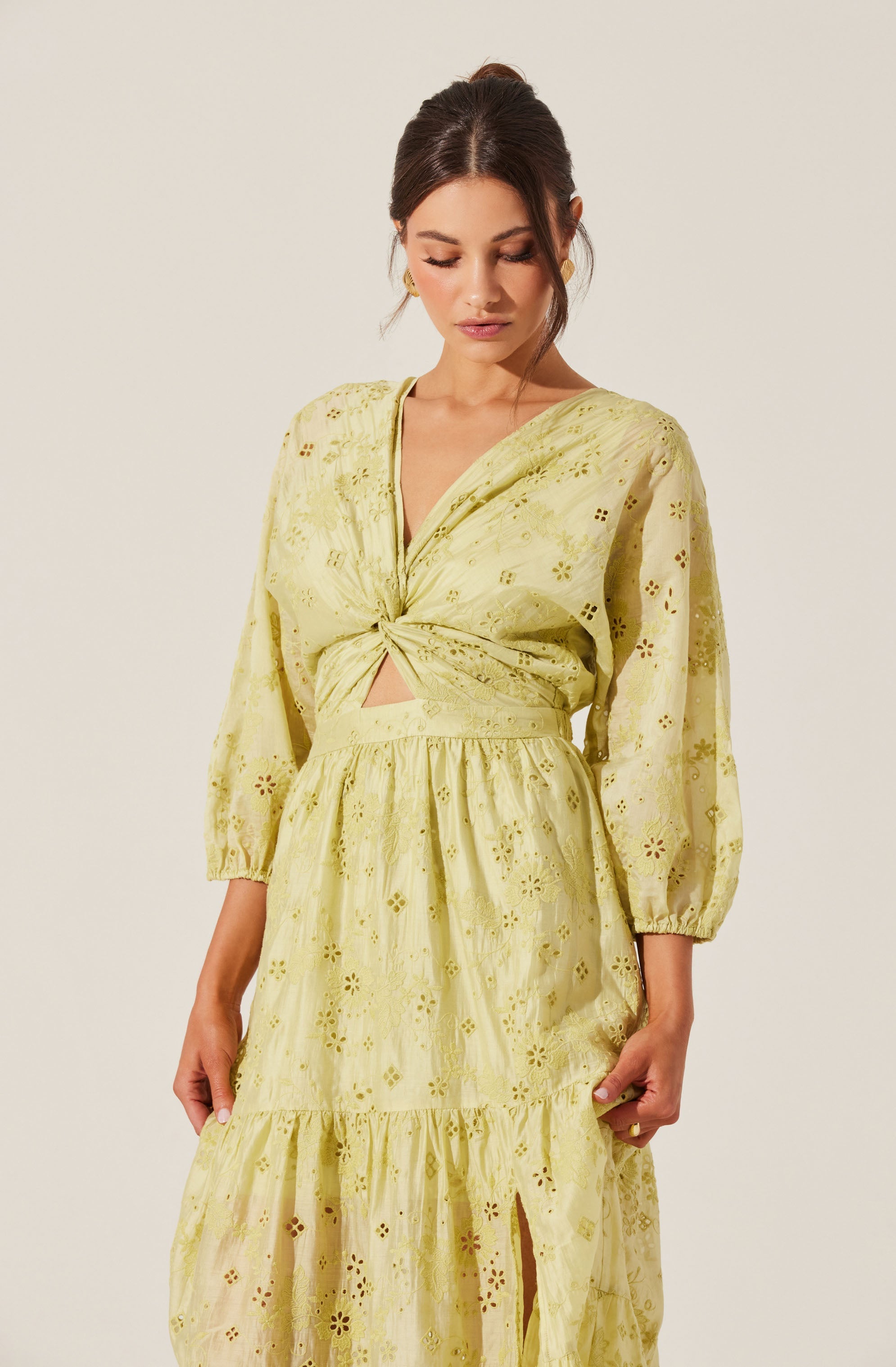 A woman is wearing a yellow, eyelet embroidered matching set consisting of a cropped, long-sleeve top with a front knot and a high-waisted, tiered skirt with a front slit. The fabric features floral and geometric cut-out patterns.