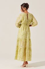 A woman is wearing a long, yellow dress made of lightweight, embroidered fabric with eyelet details. The dress features long puffed sleeves, a gathered waist, and a tiered skirt that reaches her ankles. The back of the dress has a keyhole opening tied with a small bow at the neck. She is also wearing beige high-heeled shoes and has her hair styled in an elegant updo.