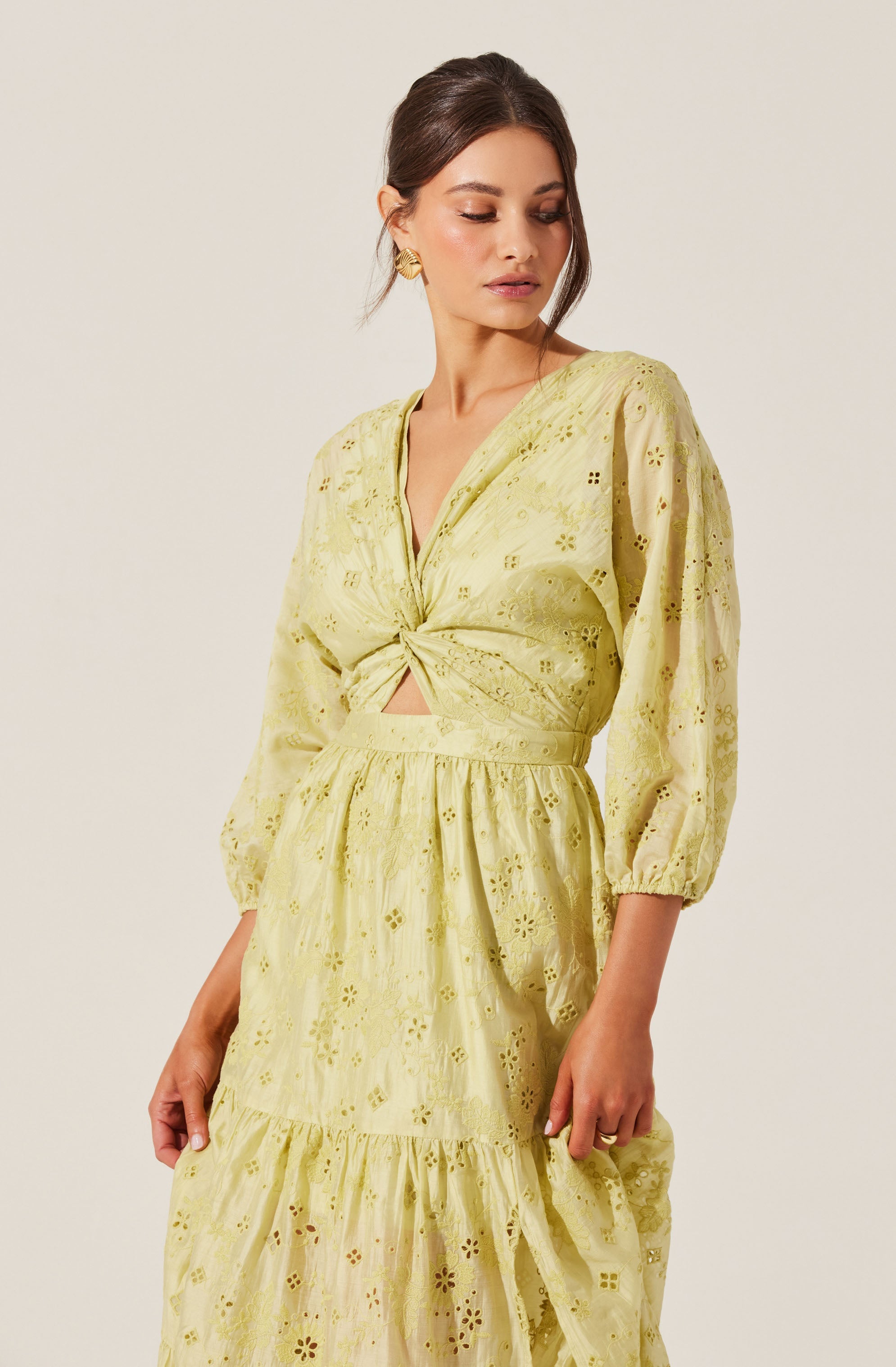 A woman is wearing a yellow eyelet lace dress with a V-neckline and long, puffed sleeves. The dress features a twist-front detail at the bust, creating a small cutout above the waist. The skirt is tiered with a ruffle at the hem, and the fabric has intricate floral patterns with eyelet cutouts throughout. She accessorizes with large, gold, geometric earrings.
