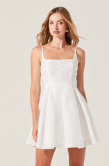 A woman is wearing a sleeveless white dress with thin shoulder straps. The dress has a textured fabric with a subtle crinkled appearance and features a fitted bodice that extends into a flared, knee-length skirt.