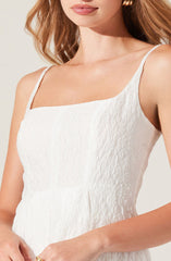 A woman is wearing a sleeveless white dress with thin spaghetti straps. The dress has a textured, crinkled fabric appearance, and features a straight neckline. The overall style is simple and elegant.