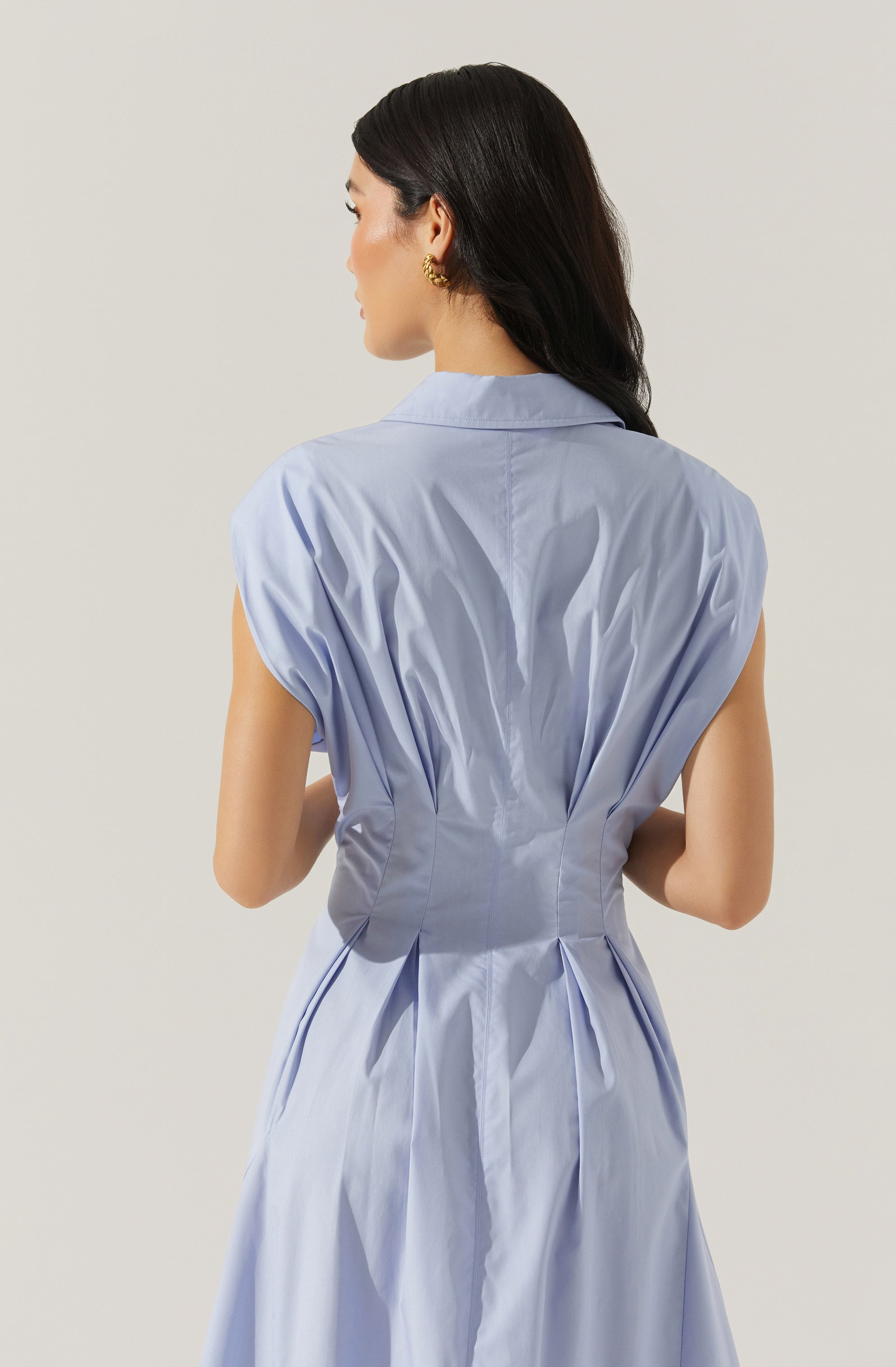 A woman is wearing a light blue, sleeveless dress with a structured waist and pleated, voluminous shoulders. The dress has a collar and a fitted silhouette that accentuates the waist, with pleats running down from the waistline.