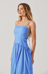 A woman is wearing a sleeveless, light blue dress with thin spaghetti straps. The dress features a fitted bodice and a gathered, flowing skirt. On the side of the dress, there is a lace-up detail with crisscrossing laces that tie into a bow on the right side. The fabric appears to be lightweight and has a slightly shiny finish.