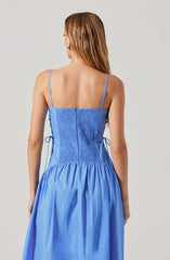 A woman is wearing a sleeveless, light blue dress with thin spaghetti straps. The dress features a fitted bodice with shirred or smocked fabric at the back, and a gathered skirt that falls to a midi length. The material appears lightweight and shiny, likely satin or silk.