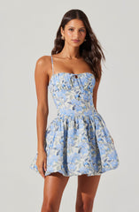 A woman is wearing a sleeveless, fitted dress with thin spaghetti straps. The dress features a square neckline with a small keyhole cutout at the center, and the fabric has a light blue and white floral pattern with hints of darker blue. The waistline is slightly cinched, and the skirt is voluminous and gathered, ending above the knee.