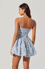 A woman is wearing a sleeveless, fitted dress with thin spaghetti straps. The dress features an elastic smocked bodice and a gathered, voluminous skirt. The fabric has a light blue background with a floral pattern in shades of blue, gray, and white.