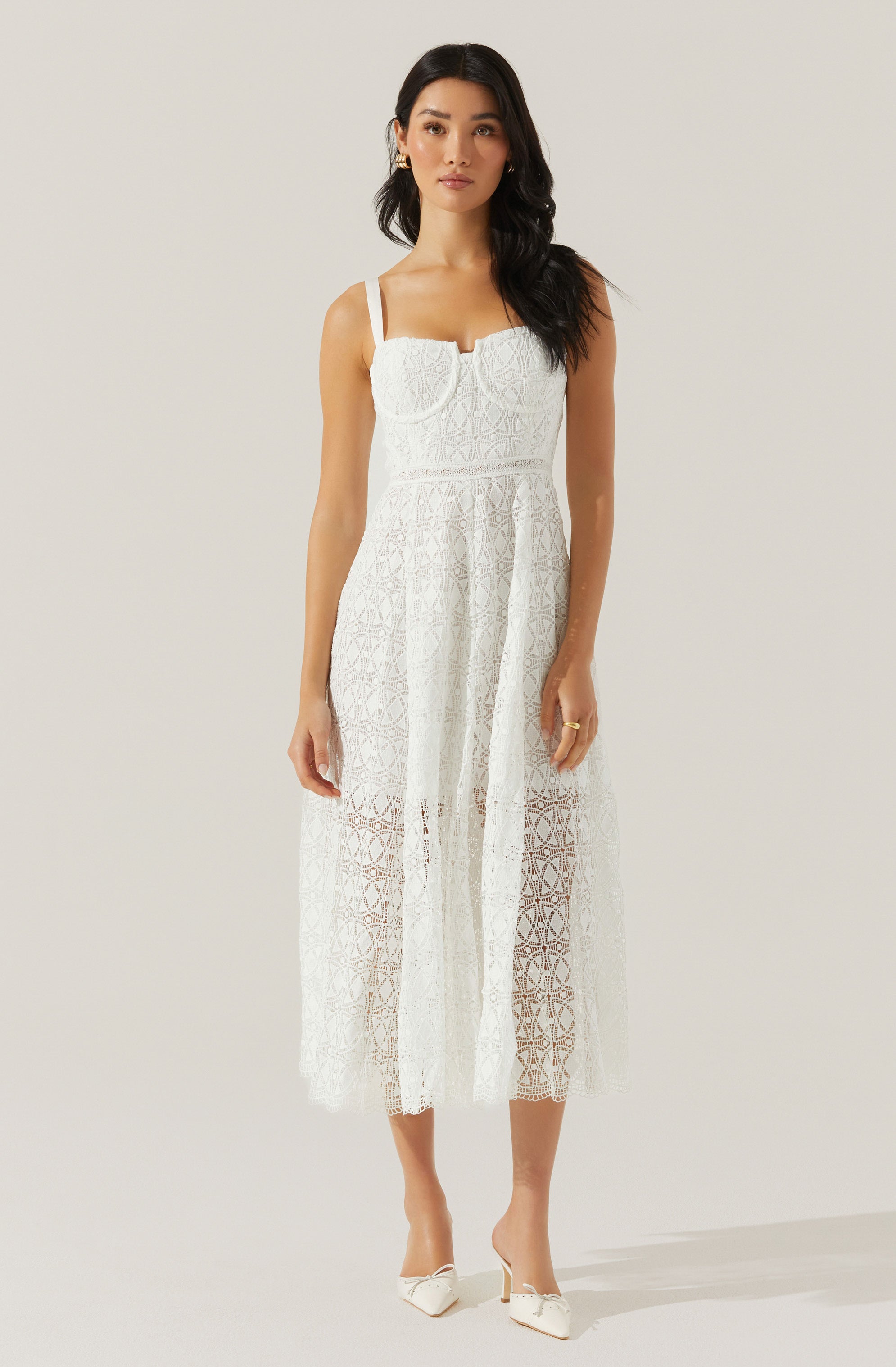 The woman is wearing a white, sleeveless, lace dress with a fitted bodice and straps over her shoulders. The dress features a sheer, patterned lace fabric that extends to mid-calf length, with a lining underneath only in the bodice area, leaving the lower part semi-transparent. She is also wearing white heeled shoes with pointed toes and decorative bows.