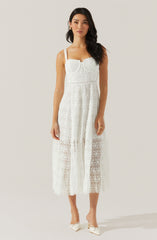 The woman is wearing a white, sleeveless, lace dress with a fitted bodice and straps over her shoulders. The dress features a sheer, patterned lace fabric that extends to mid-calf length, with a lining underneath only in the bodice area, leaving the lower part semi-transparent. She is also wearing white heeled shoes with pointed toes and decorative bows.