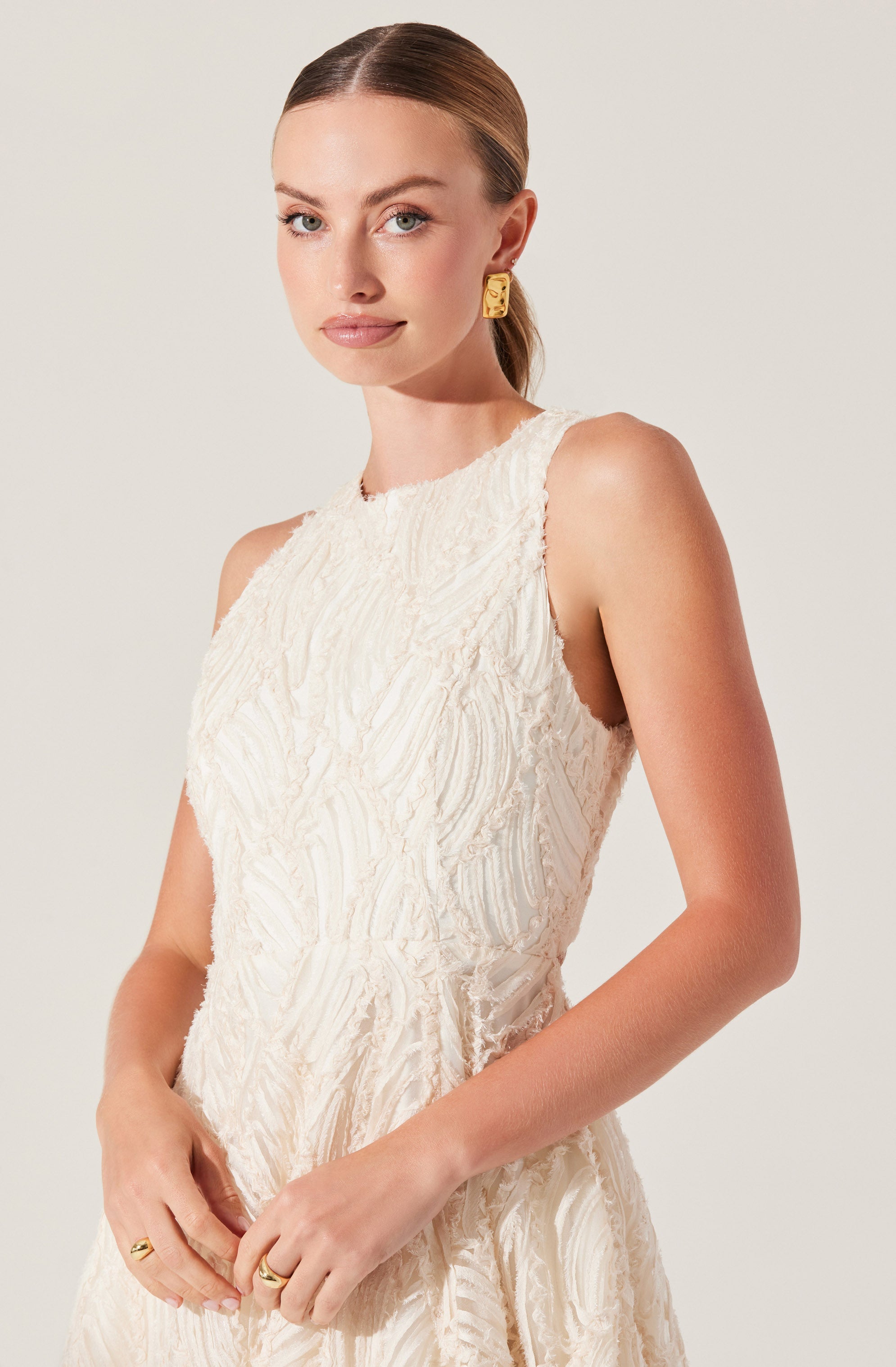 A woman is wearing a sleeveless, textured, cream-colored dress with a high neckline. She has her hair pulled back and is accessorized with large, gold, geometric earrings and rings on her fingers.