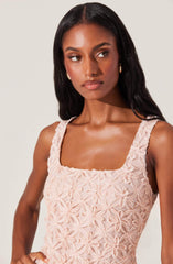 A woman with dark, wavy hair is wearing a sleeveless, light pink dress with a textured floral pattern made of fabric loops. The dress has a square neckline and wide shoulder straps. She is accessorized with small, gold hoop earrings.