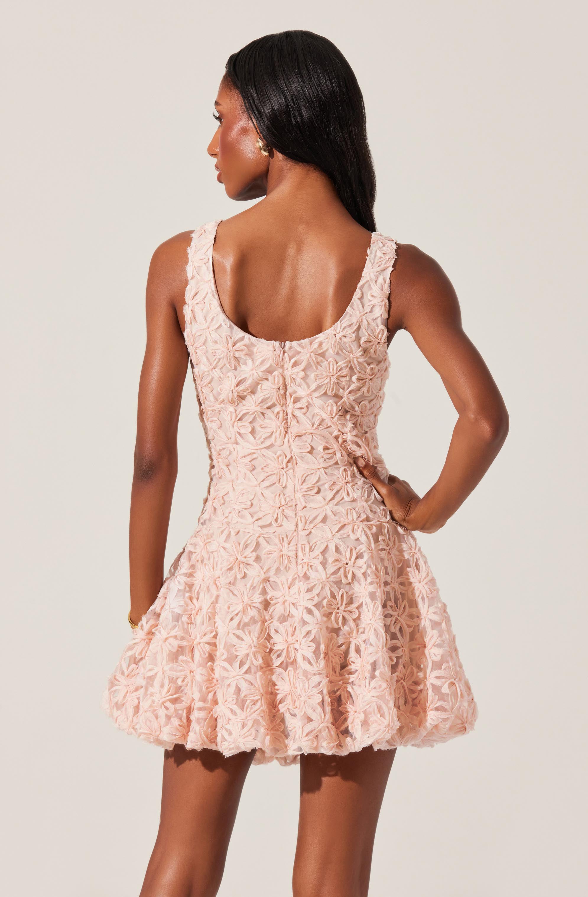 A woman is wearing a sleeveless, peach-colored dress with intricate floral embroidery. The dress has a fitted bodice with a scoop neckline and a full, flared skirt that ends above the knees. The fabric appears textured with a three-dimensional floral pattern.