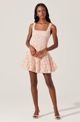 A woman is wearing a sleeveless, light pink dress with a textured floral lace overlay. The dress has wide shoulder straps, a fitted bodice, and a flared skirt that falls above the knees. She is accessorized with gold earrings, a gold bracelet, and a gold ring, and is wearing matching light pink open-toe heels.