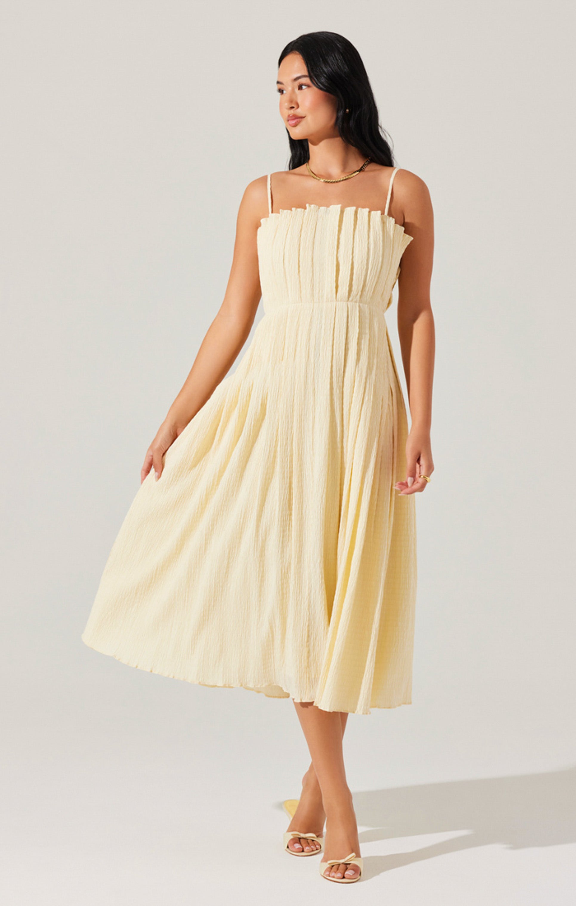 A woman is wearing a sleeveless, light yellow dress with thin spaghetti straps. The dress has a textured, pleated fabric that creates a ruffled effect around the neckline and flows down to a midi length. She is also wearing open-toe, high-heeled sandals in a matching light yellow color.