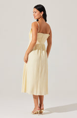 A woman is wearing a light yellow, sleeveless, midi-length dress with thin spaghetti straps and a gathered, elasticized waist. The fabric has a slightly textured, crinkled appearance. She is paired with nude high-heeled sandals.