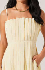 A woman wearing a sleeveless, light yellow dress with a textured, crinkled fabric. The dress has thin spaghetti straps and a gathered, ruffled bodice. She accessorized with a gold chain necklace.